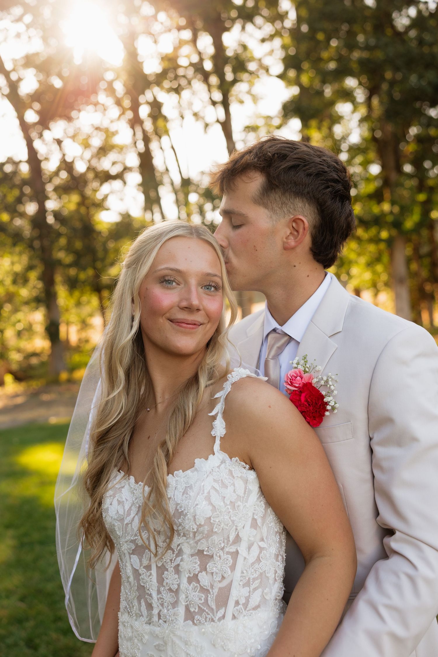 Groom tenderly kissing blonde bride on cheek in golden sunlight among trees on wedding day.