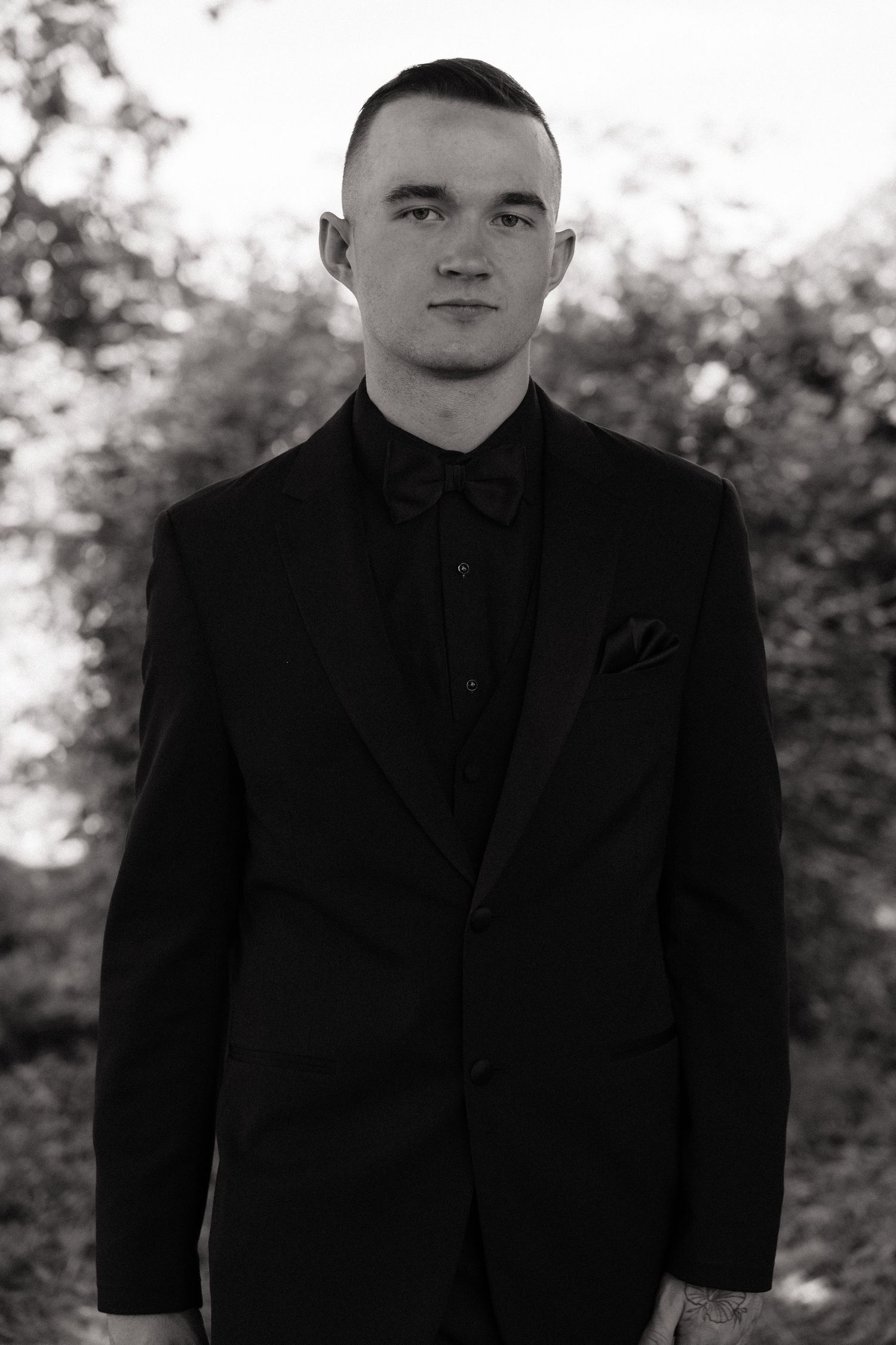Black and white portrait of a groom in a black suit with bow tie and pocket square outdoors.