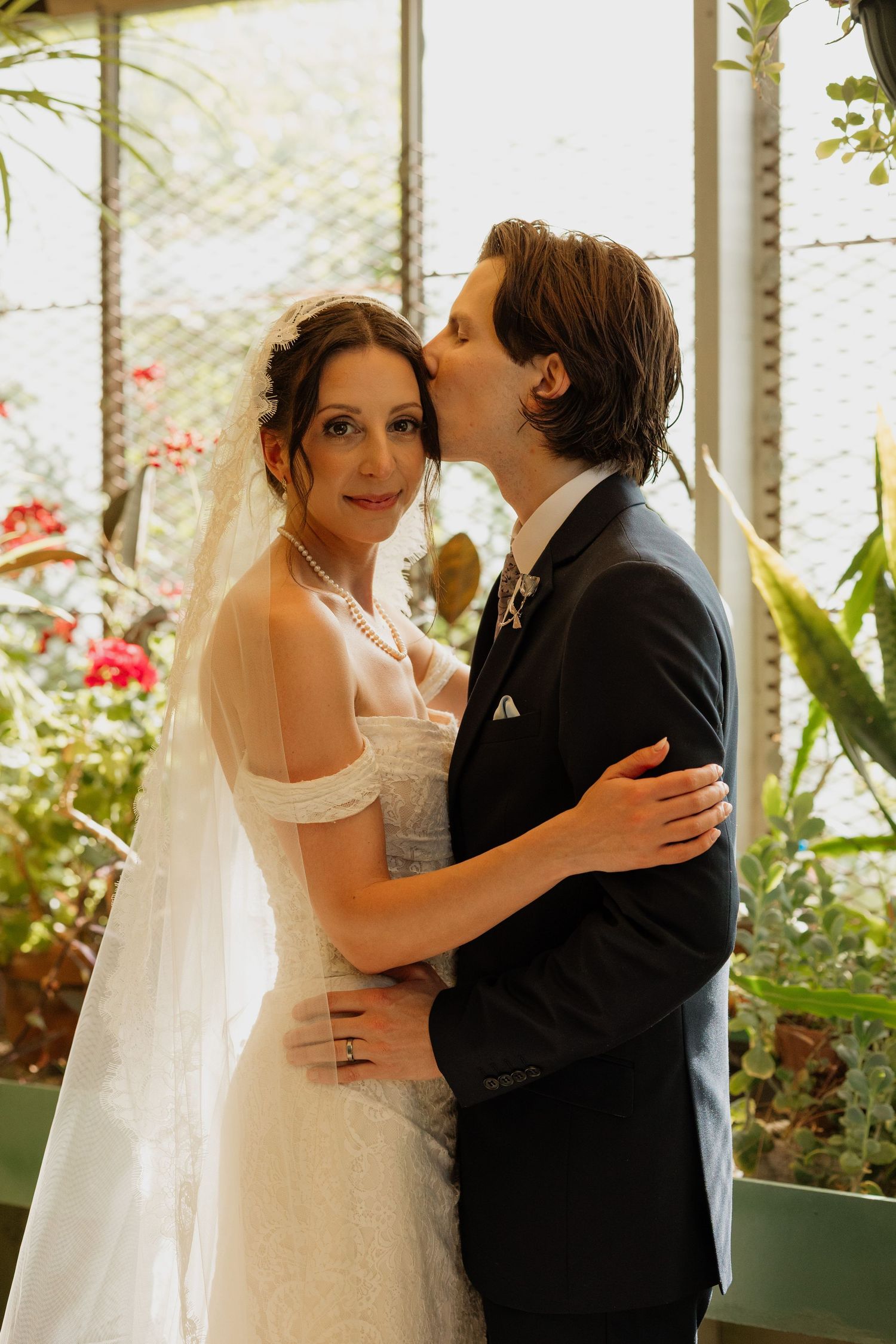 Groom in black suit kissing bride on cheek, bride wearing off-shoulder lace gown and veil in greenhouse.