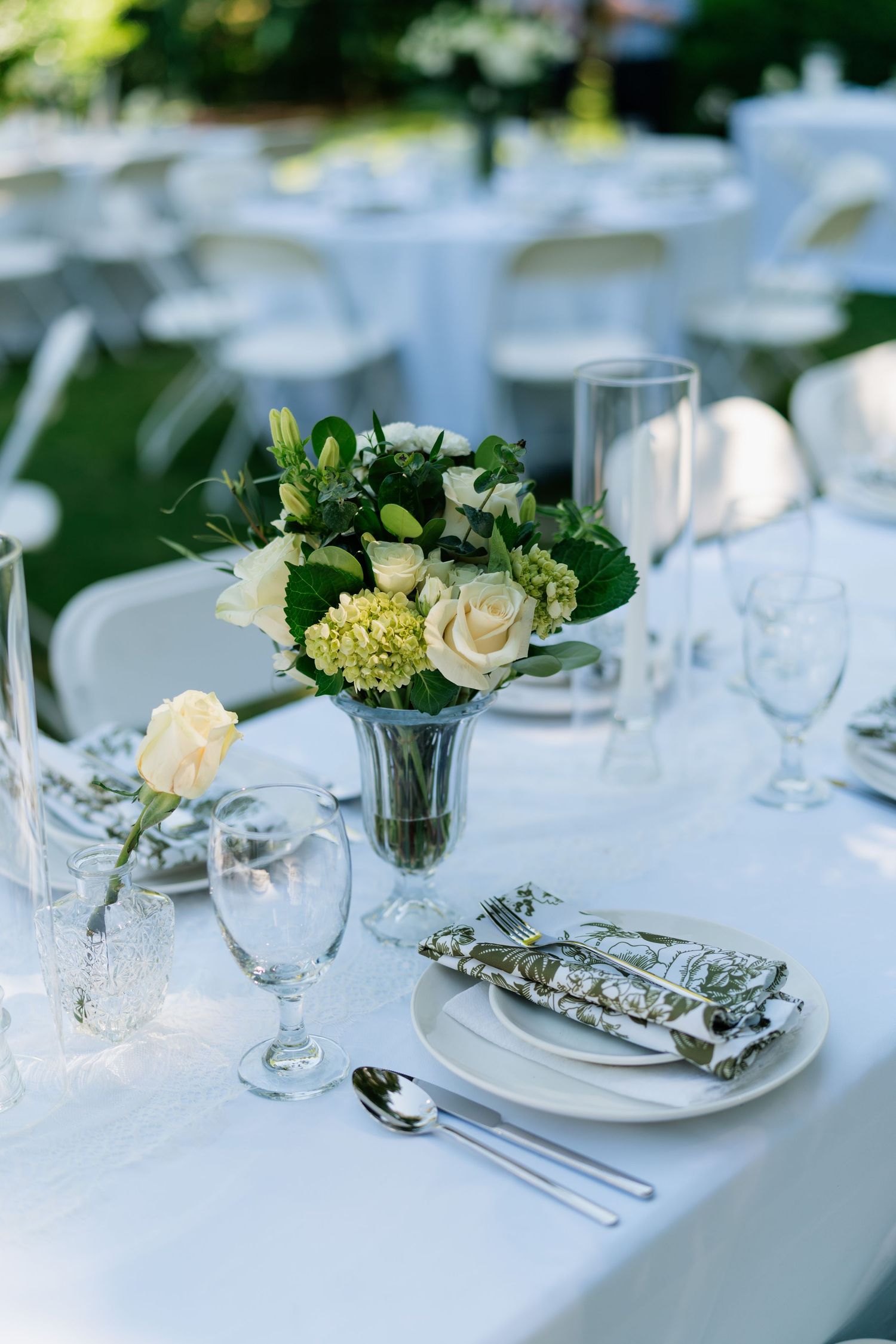 Close-up of outdoor wedding table setting with white roses, hydrangeas, crystal glassware, and patterned napkins on white linen.