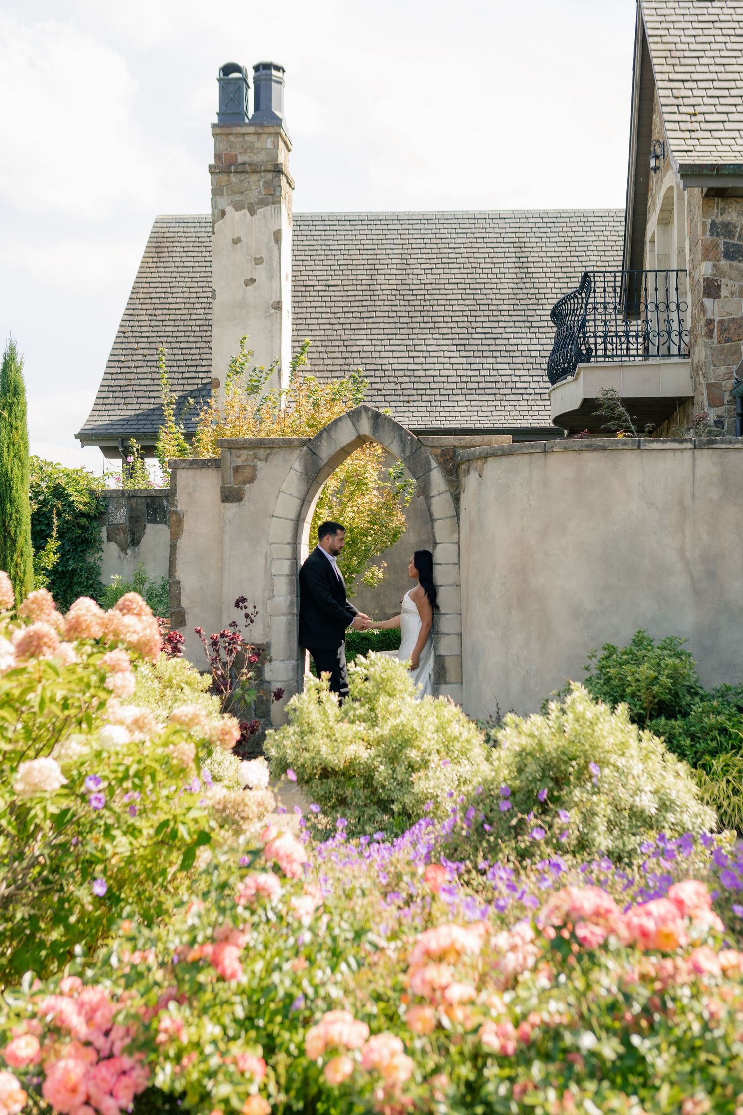 Pink and white flowers bloom in a garden against a backdrop of stone buildings with grey rooftops.