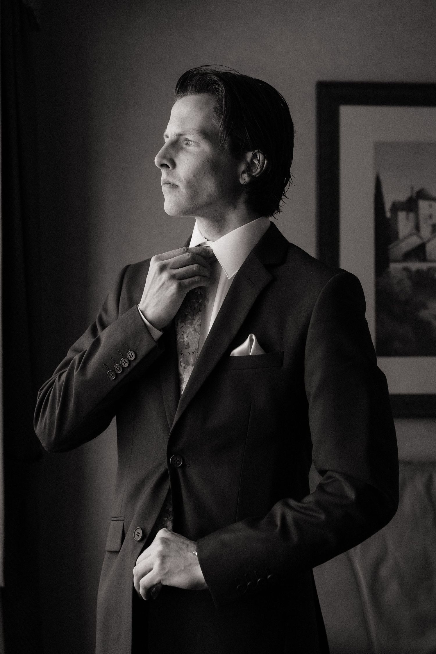 Elegant groom in black suit adjusting tie in black and white portrait photo.