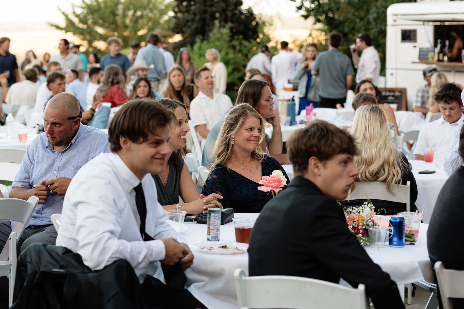 Guests in formal attire seated at white-clothed tables during an elegant outdoor wedding reception at sunset.
