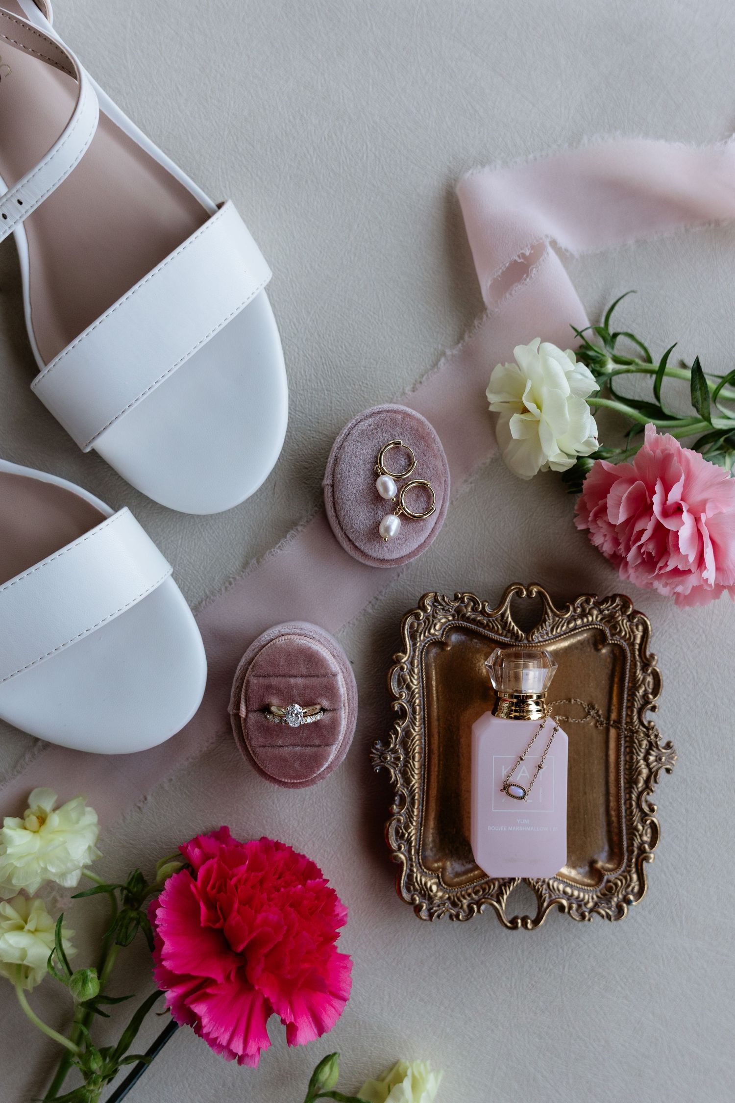 Wedding flat lay featuring white sandals, engagement ring, pearl earrings, perfume bottle, and pink flowers.