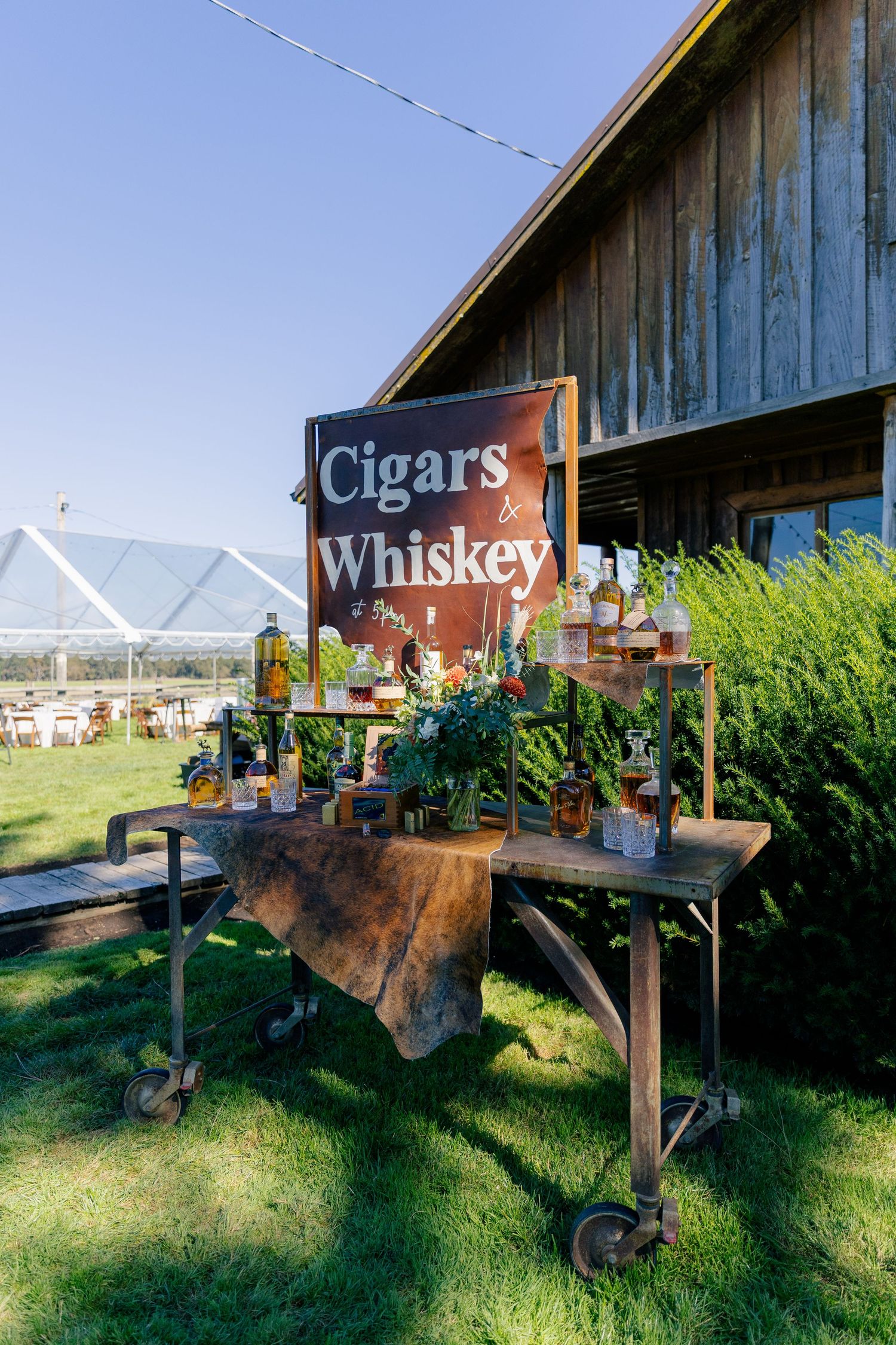 Rustic outdoor bar setup with vintage 'Cigars & Whiskey' sign and plants displayed on wooden cart against barn backdrop.
