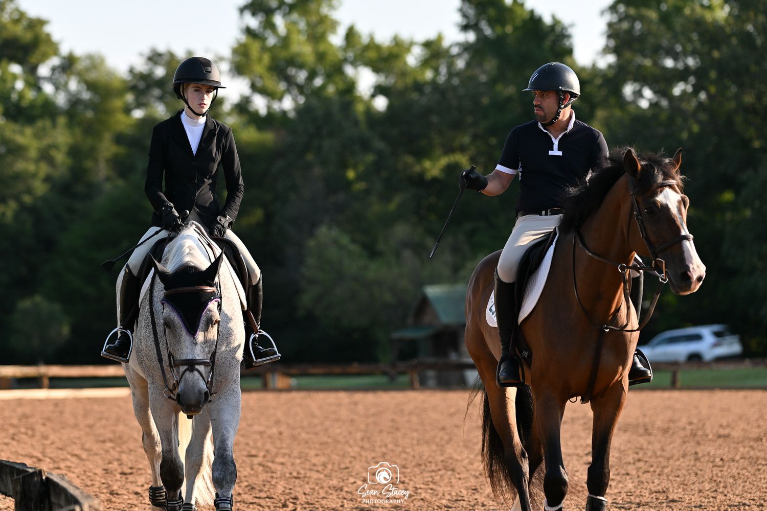 Two equestrians in formal riding attire practicing on horses in a sandy arena during evening light.