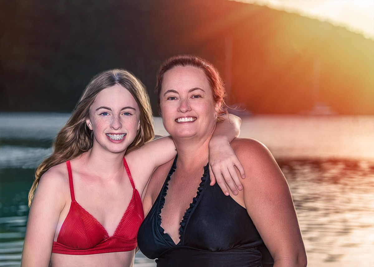 Two friends in swimwear smile together at sunset by a lake with golden sunlight glowing in the background.