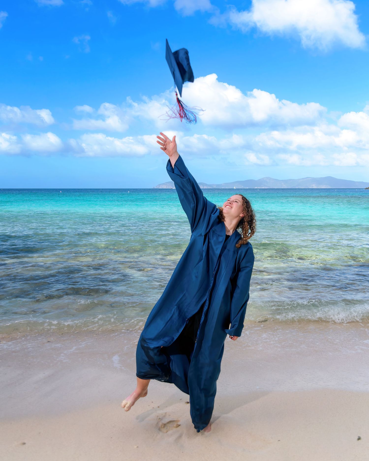 A graduate in a navy blue gown celebrates on a tropical beach with crystal clear turquoise waters and blue skies.