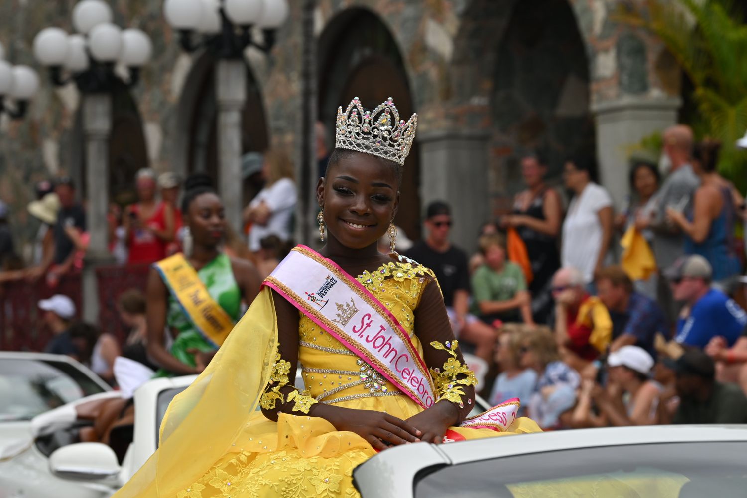 Pageant participants in colorful dresses and crowns wave to spectators during a parade celebration.