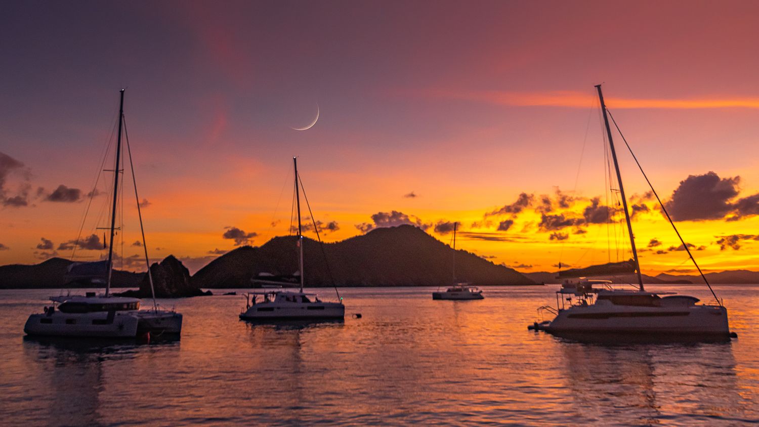 Sailboats anchored in calm waters during a vibrant orange and purple Caribbean sunset.