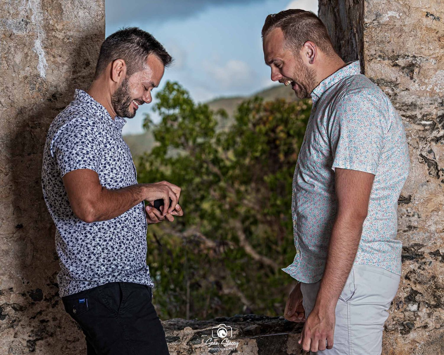 A couple in casual attire embraces against a rustic stone wall in a romantic outdoor setting.
