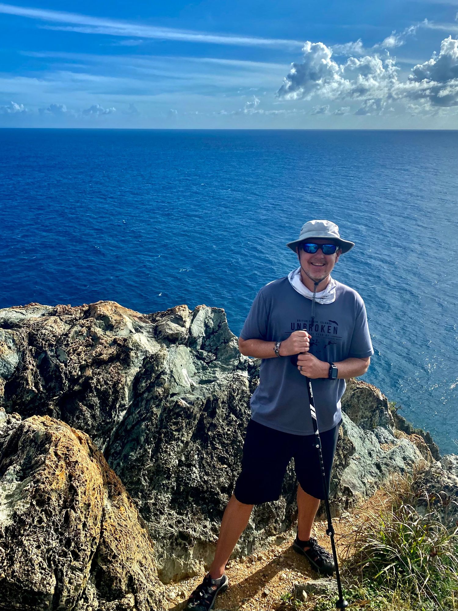 Hiker with hiking poles stands on rocky coastal cliff overlooking deep blue ocean waters on sunny day with white clouds.
