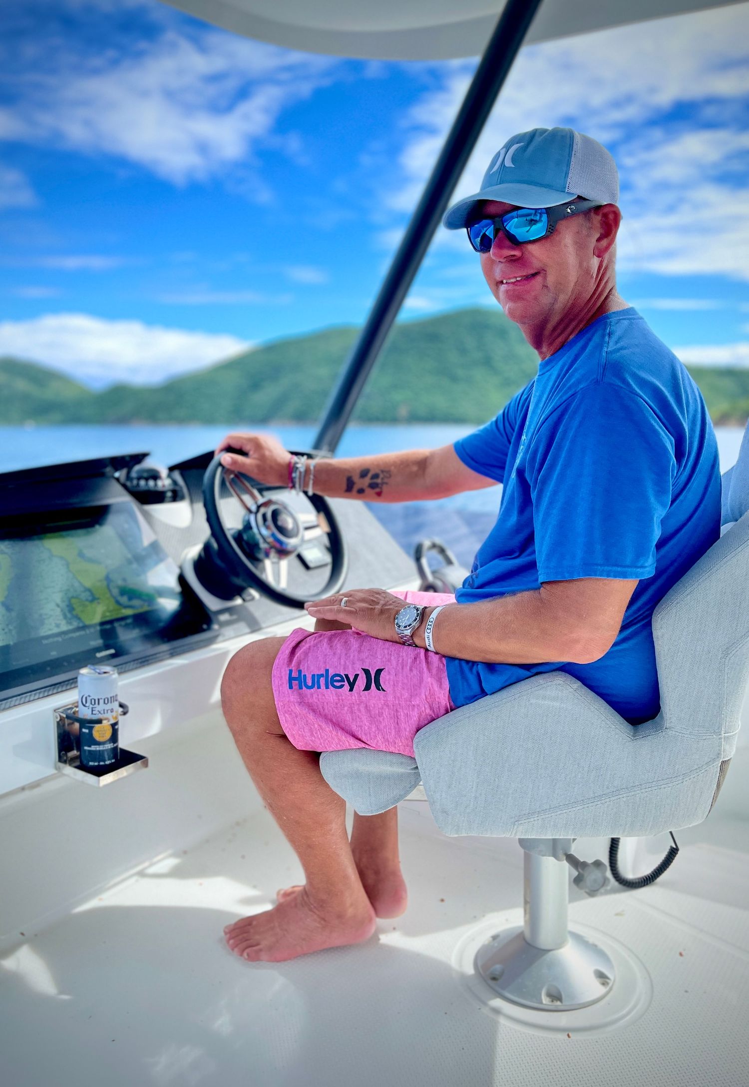Person in blue shirt operating boat controls while seated at helm station under bright blue sky with mountains visible.