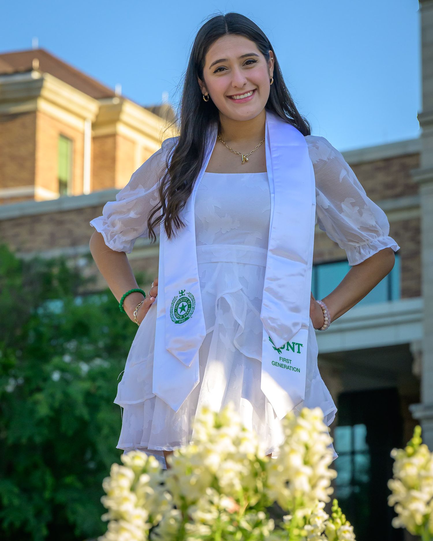 Senior in white graduation sash and dress stands smiling outdoors near yellow flowers and brick building on the campus of UNT.