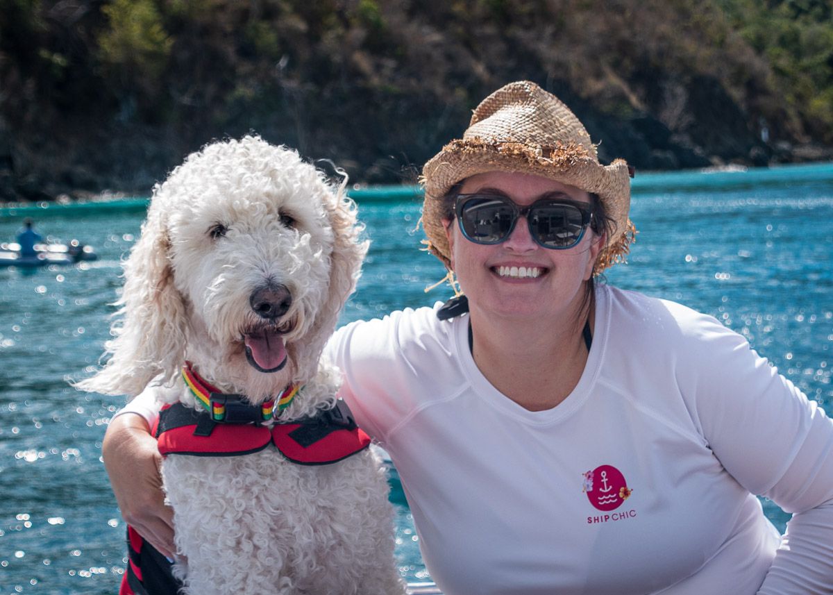 A happy Goldendoodle dog and someone in a white shirt enjoy a sunny day on a boat with blue water in the background.