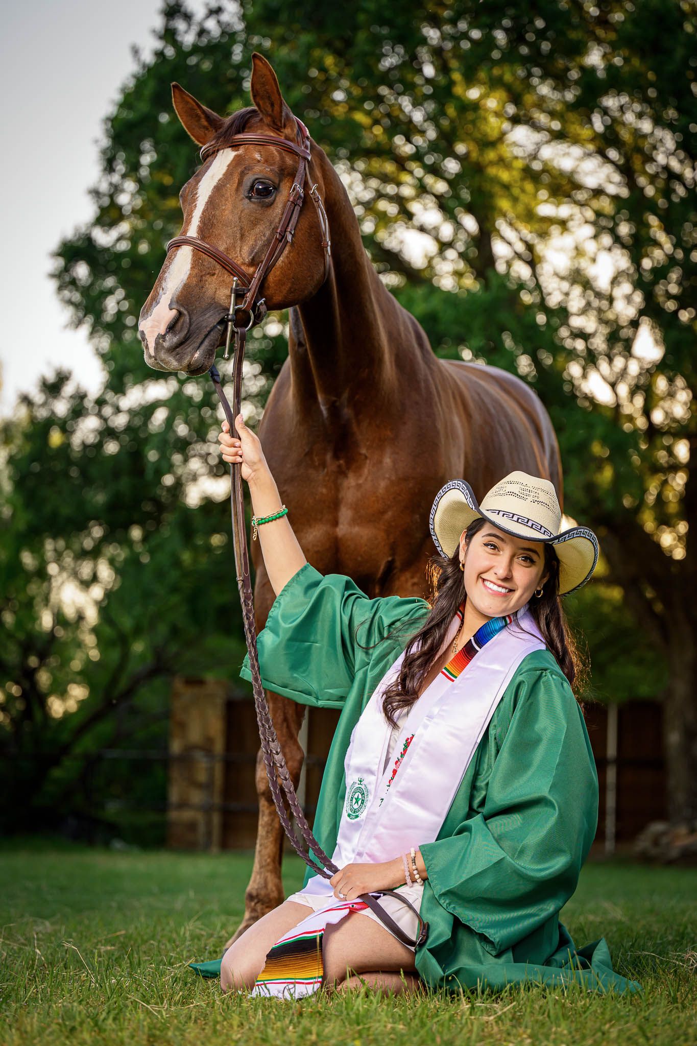 A brown horse and rider in a green jacket relax on grassy ground during golden hour.