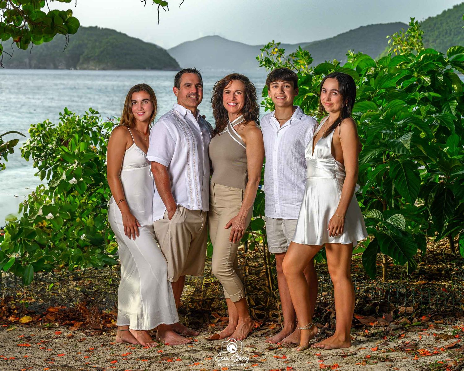 A group of five people in light summer attire pose together on a tropical beach with mountains and ocean in the background.