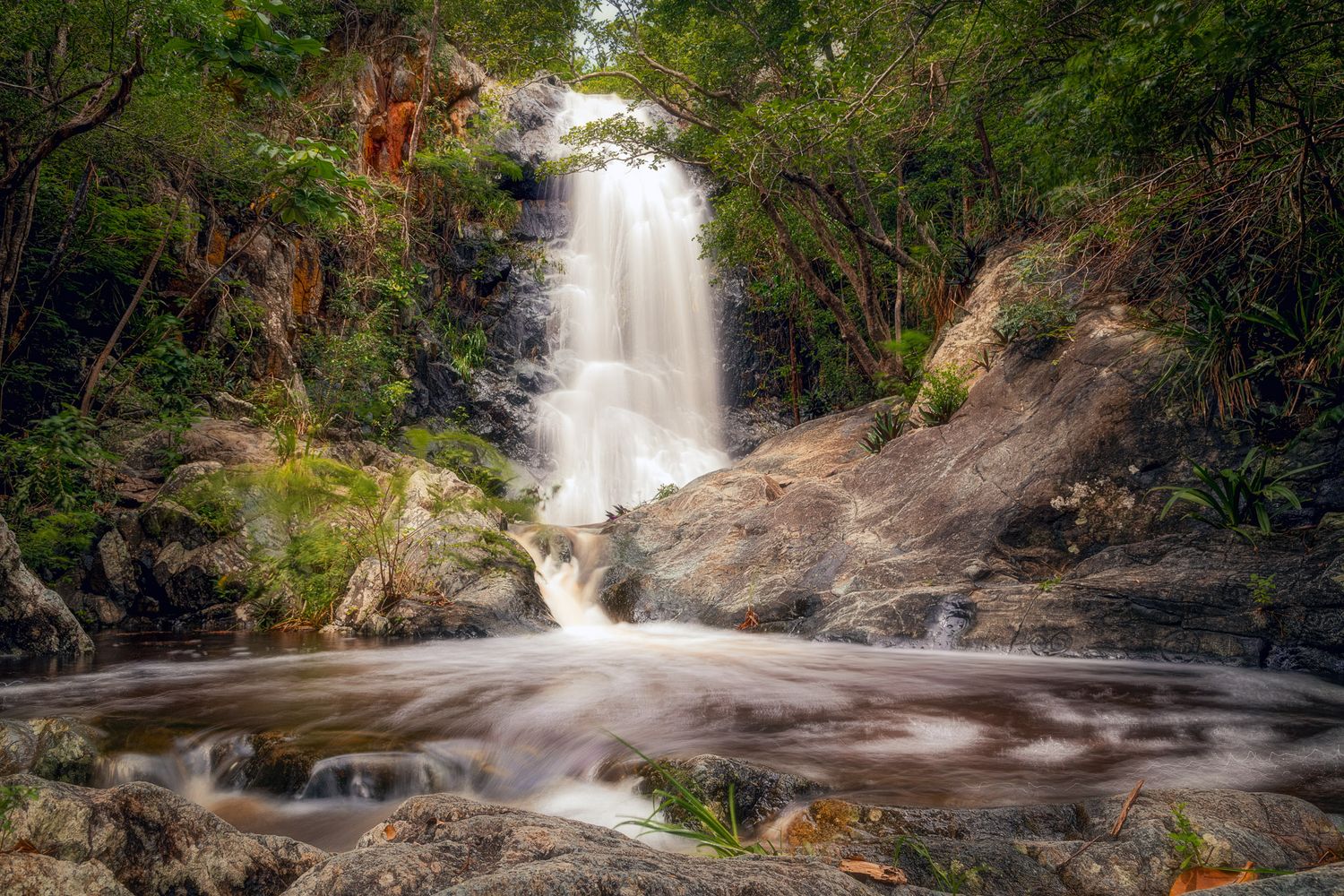 A series of cascading waterfalls flowing through lush tropical forest with rocky outcrops and green vegetation.