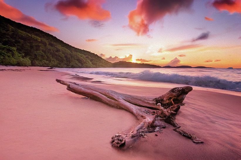 Driftwood rests on pink sand beach at sunset with dramatic red clouds and rolling waves in Hawaii.