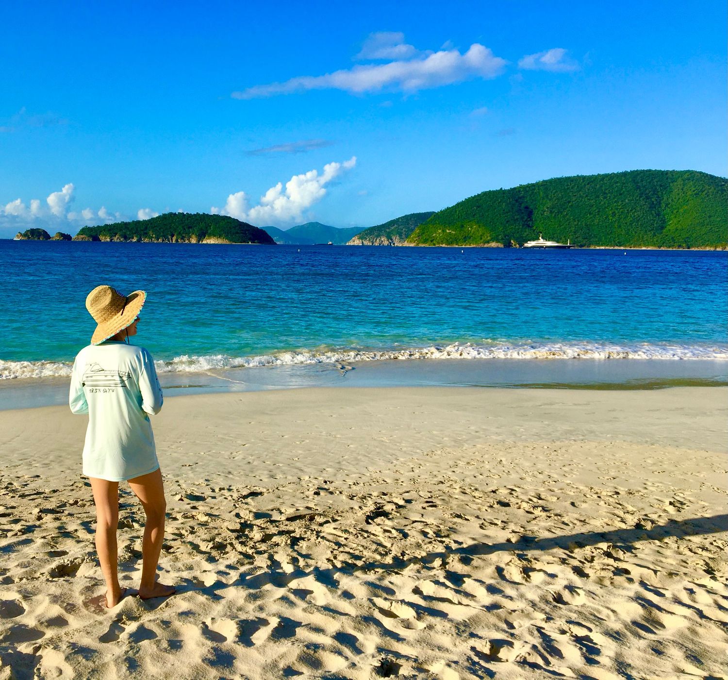 A person in a sun hat and coverup stands on a sandy beach with turquoise waters and green hills in the background.