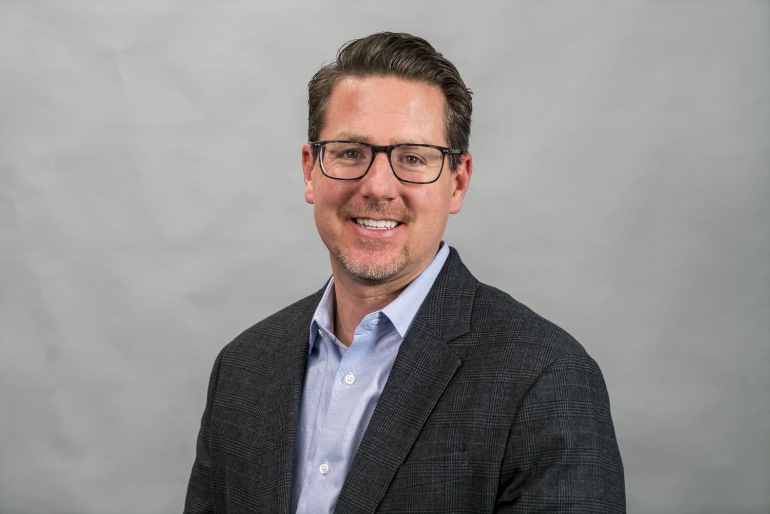 Professional headshot portrait of someone in a dark suit jacket and white dress shirt against a gray background.