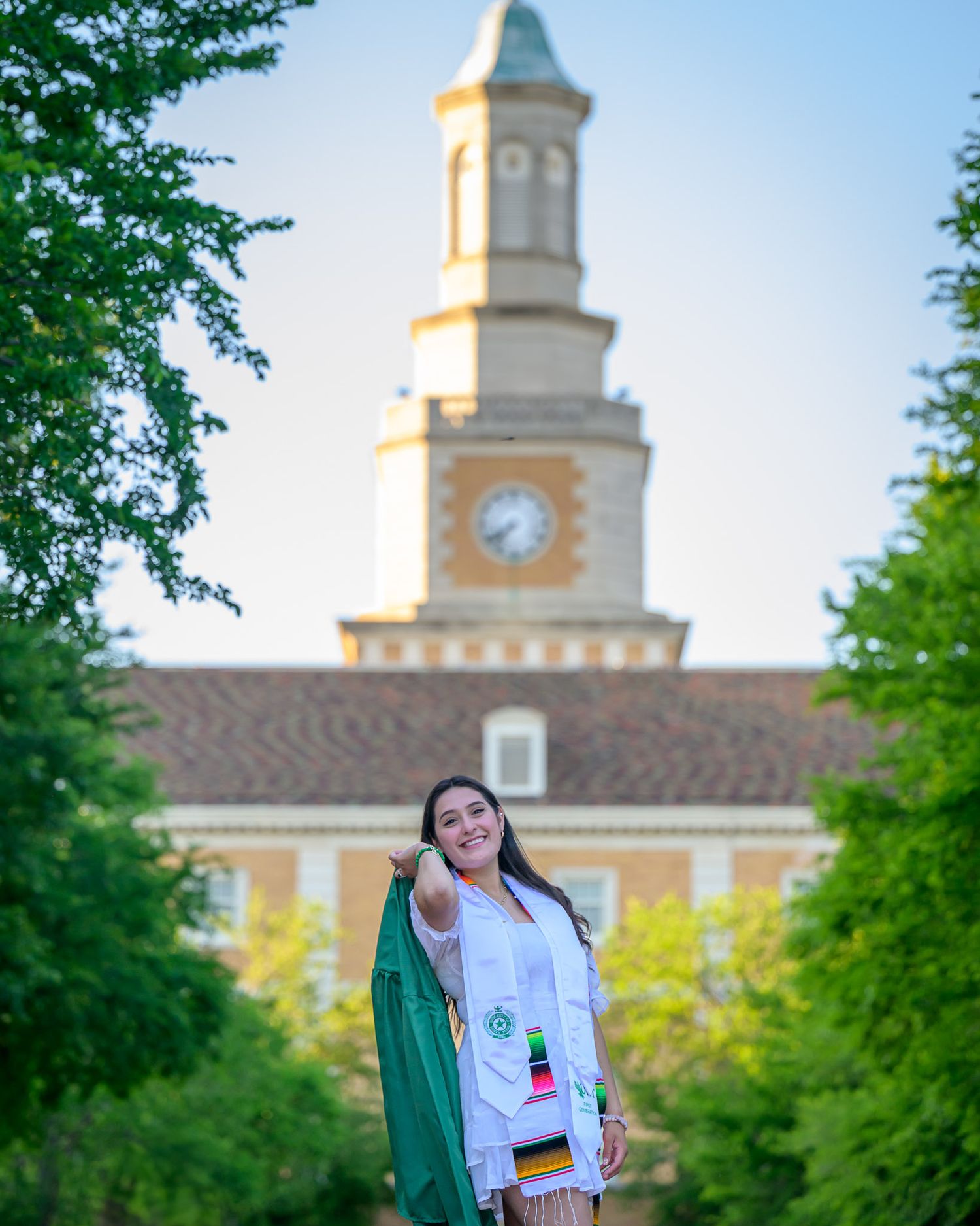 UNT senior girl graduate stands on a college campus path with a historic clock tower building in the background during golden hour.