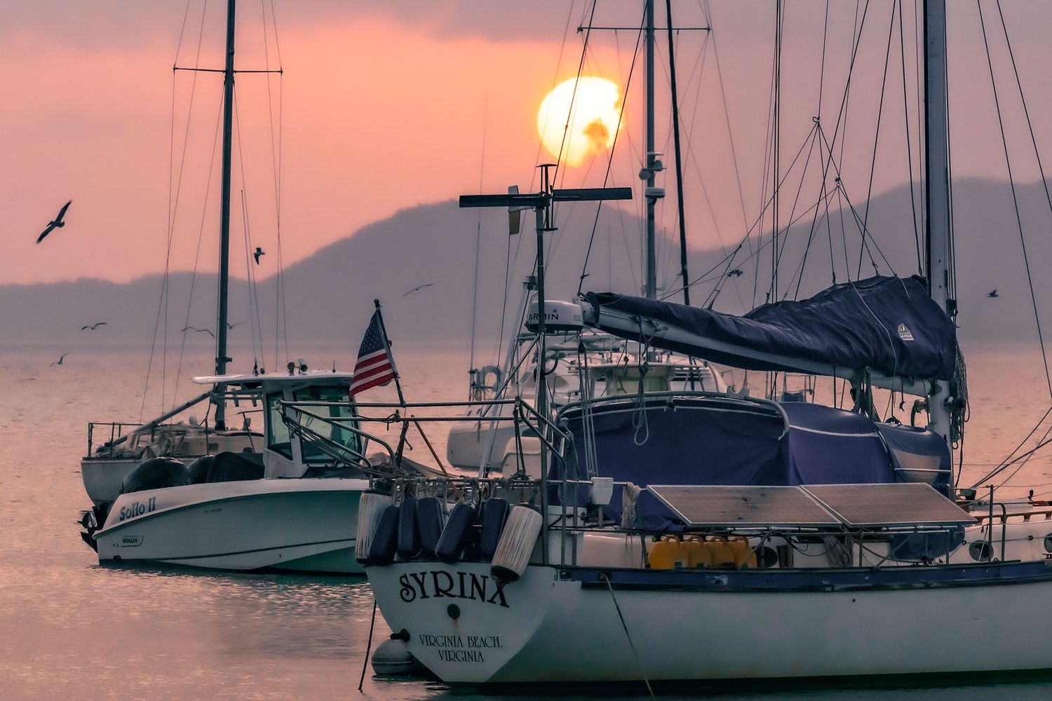 Sailboats anchored in a harbor at sunset with a golden sun setting behind mountains over calm waters.