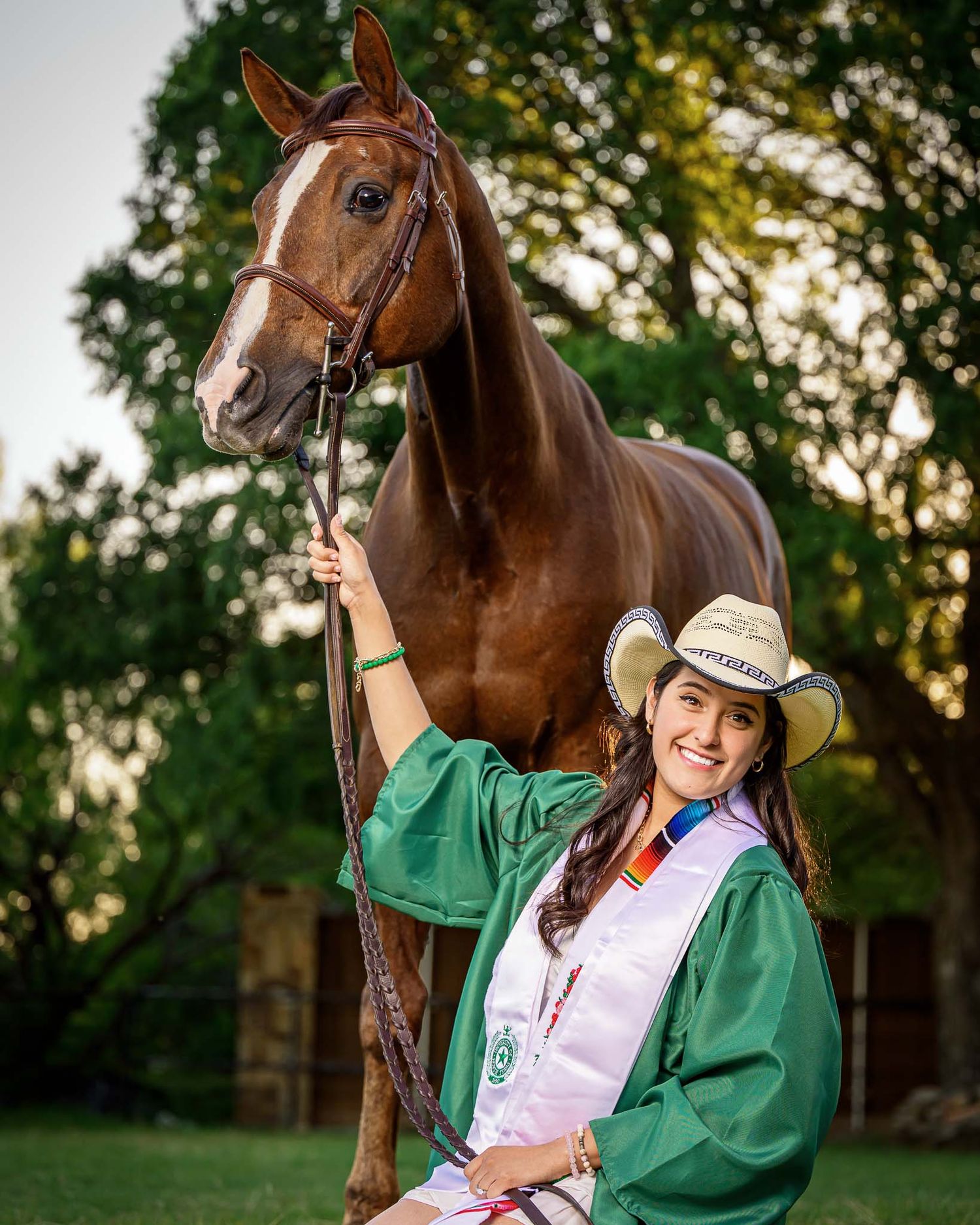 College graduate poses with a brown horse wearing a green graduation gown and white dress in an outdoor setting taken by Sean Stacey.