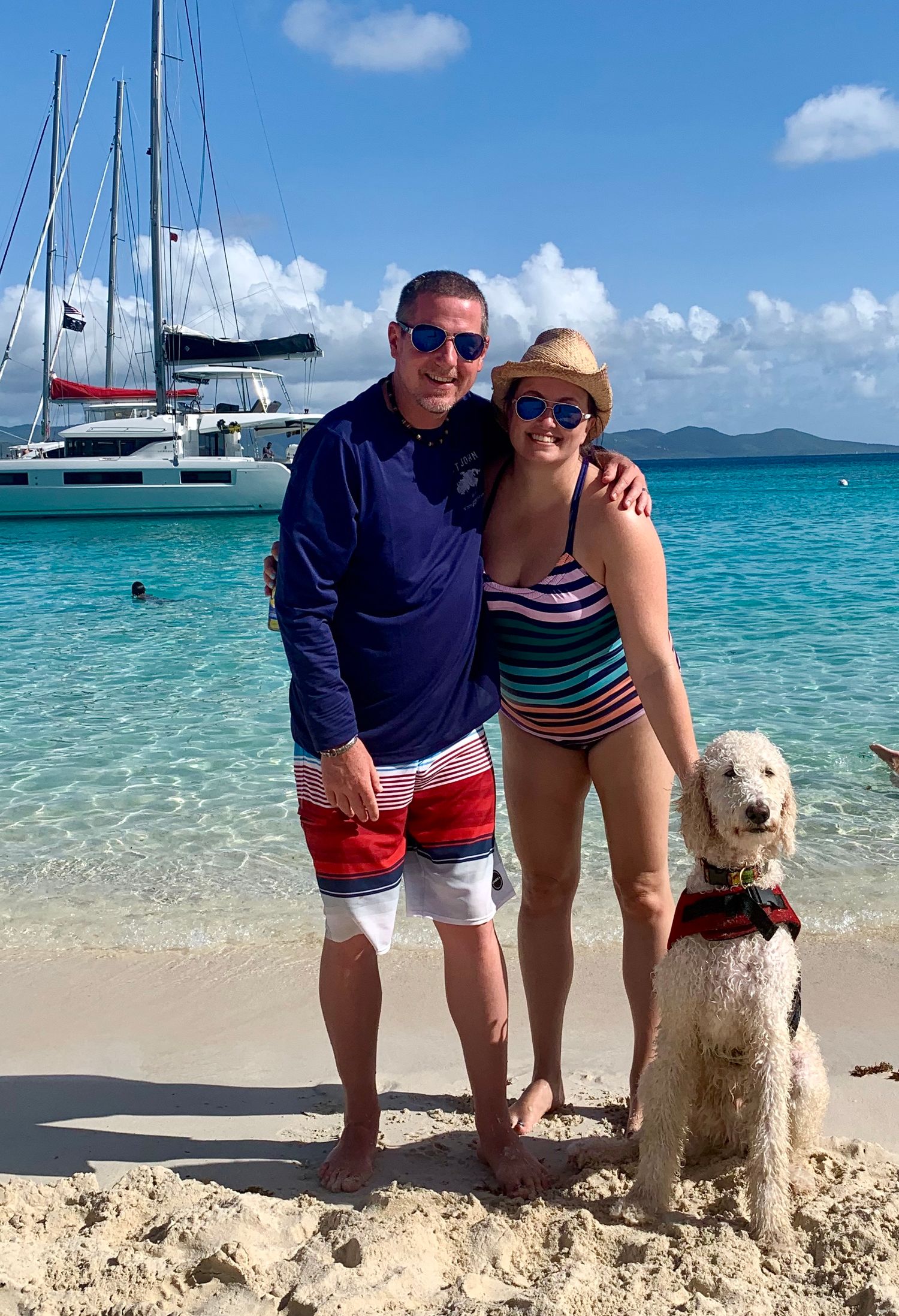 A white dog and companions enjoying a sunny day on a pristine beach with turquoise waters and boats in background.