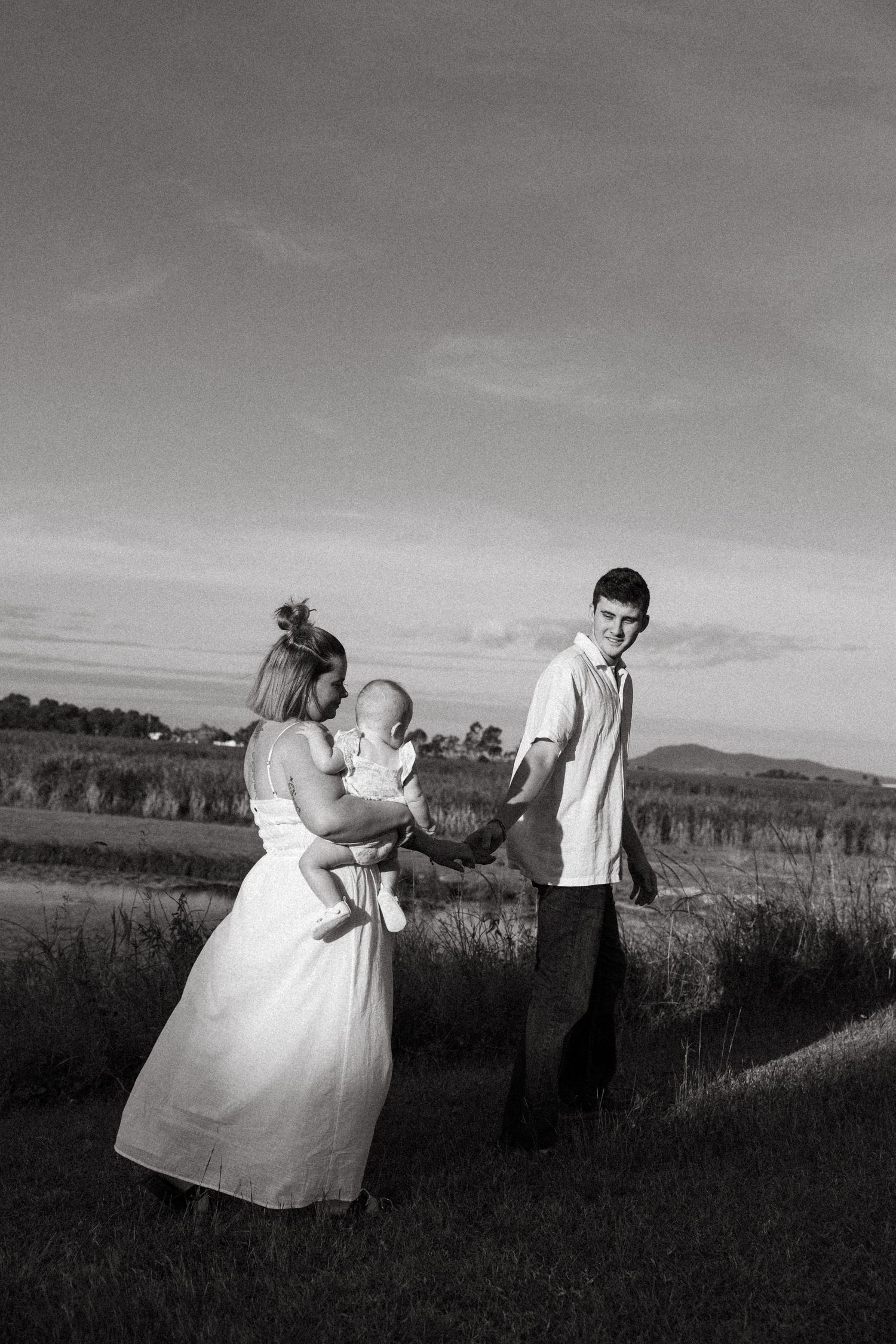 Black and white photograph of a couple in a field holding an infant against a sunset landscape backdrop.