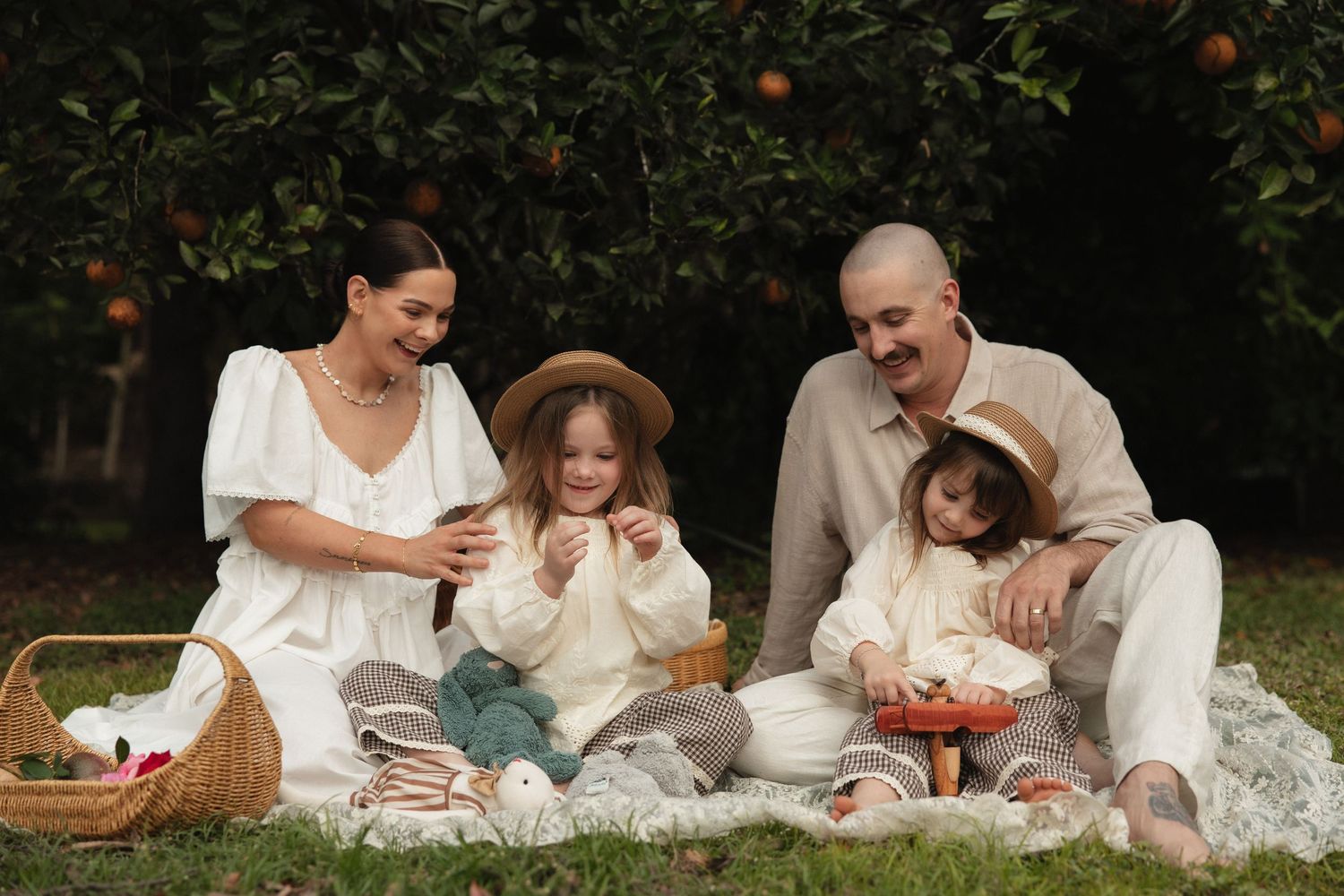 A family enjoys a picnic together on a white blanket in a sunlit garden with oranges and fruit.
