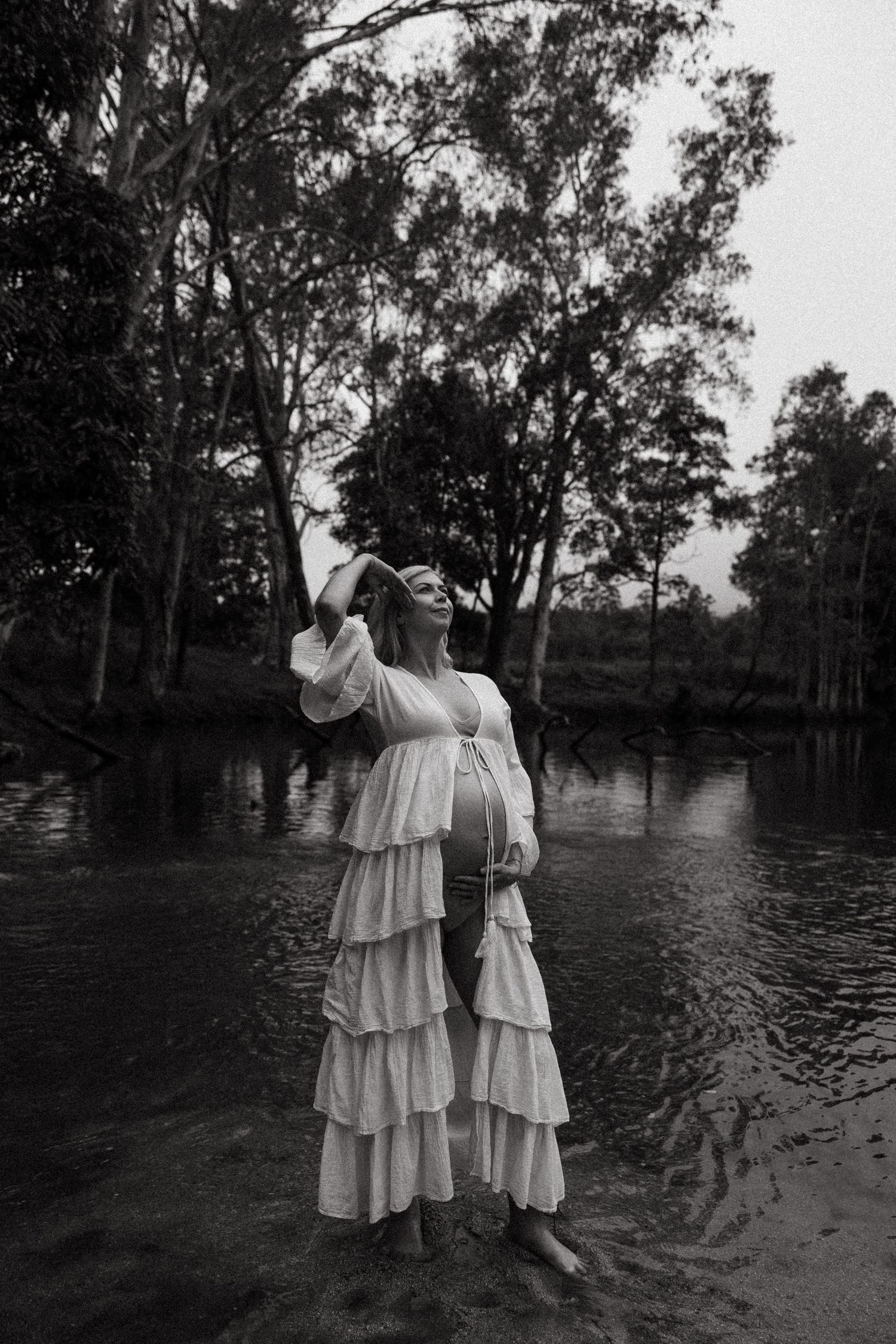 A person in a flowing white tiered dress poses gracefully by a serene pond surrounded by trees in black and white.