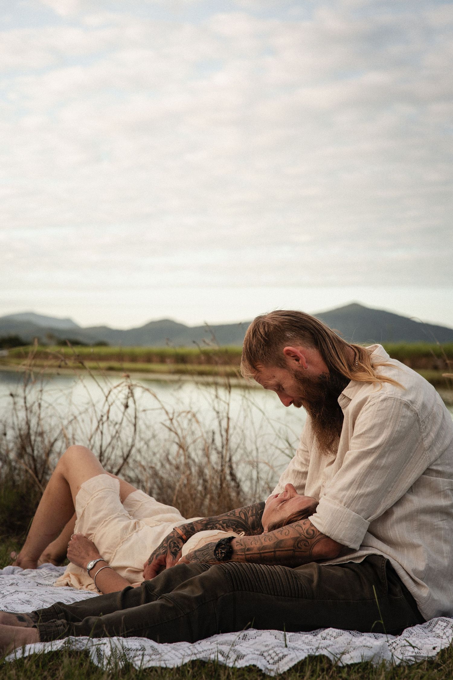 Two people having a romantic picnic by a mountain lake at sunset on a white blanket.
