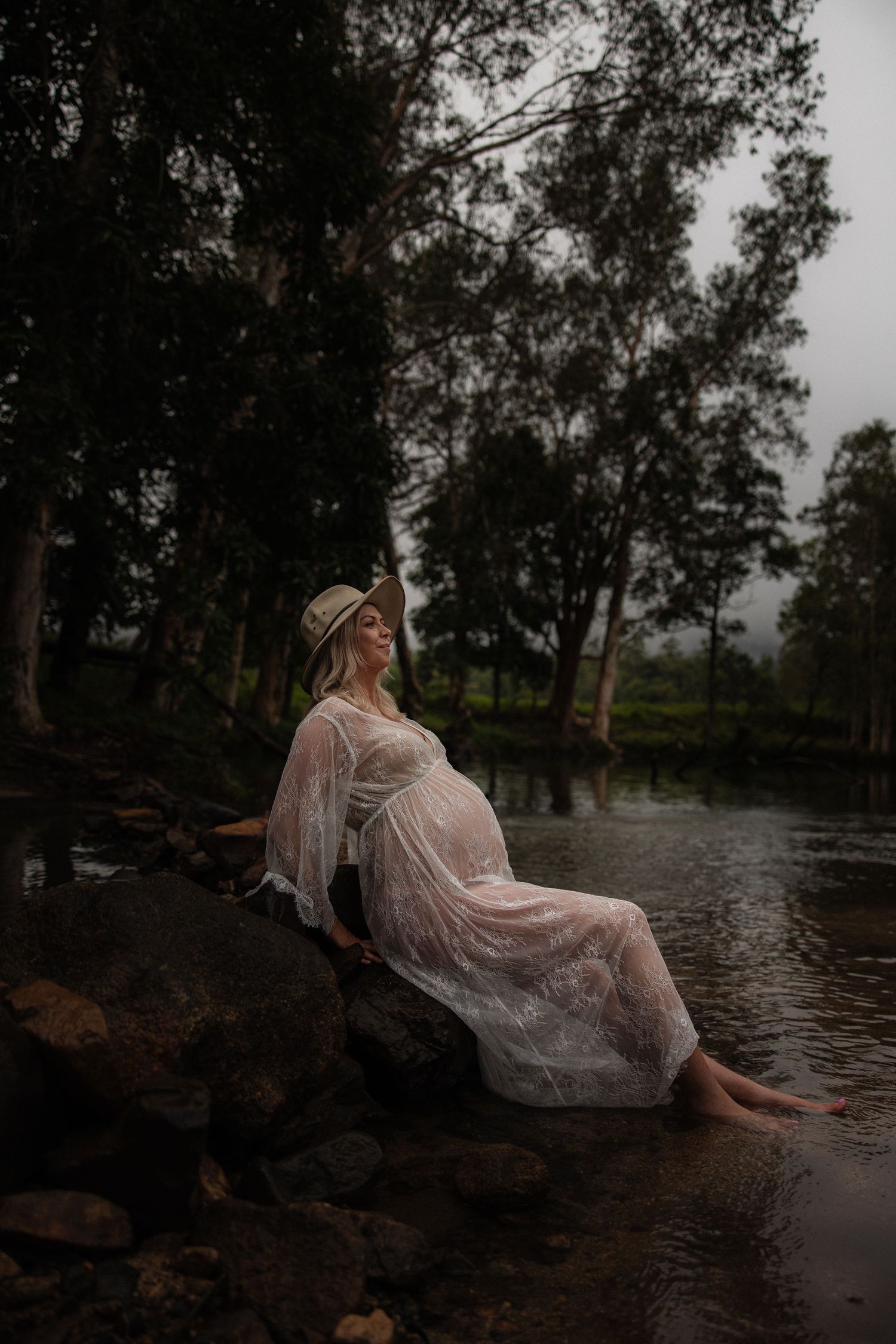 An ethereal maternity photo of a woman in a flowing white dress reclining on rocks by a river in a misty forest.