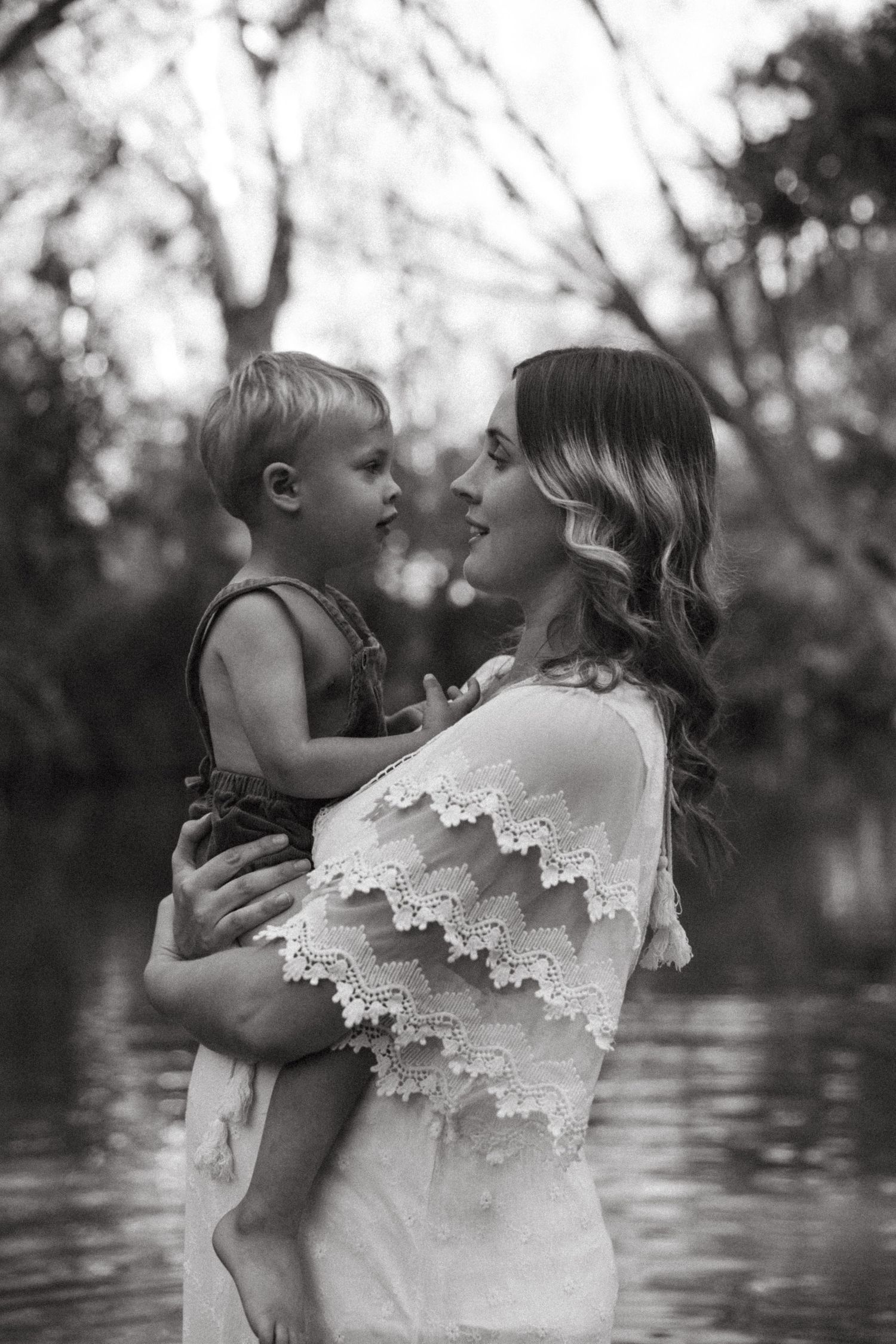 A tender black and white portrait showing a mother in a lace-trimmed dress holding a small child by a lake.