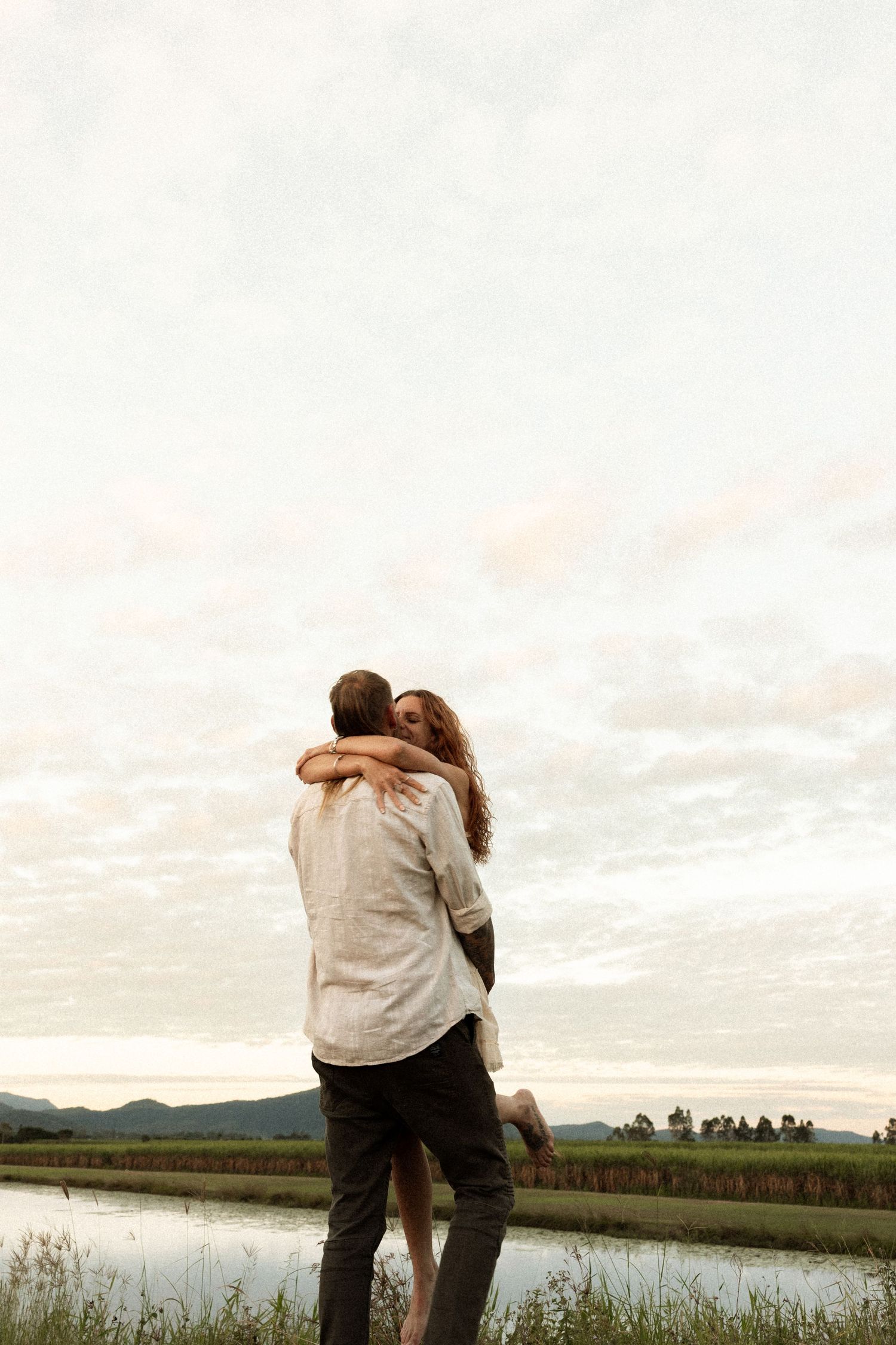 Romantic couple embraces while standing near a peaceful pond with rolling hills and soft sunset sky in background.