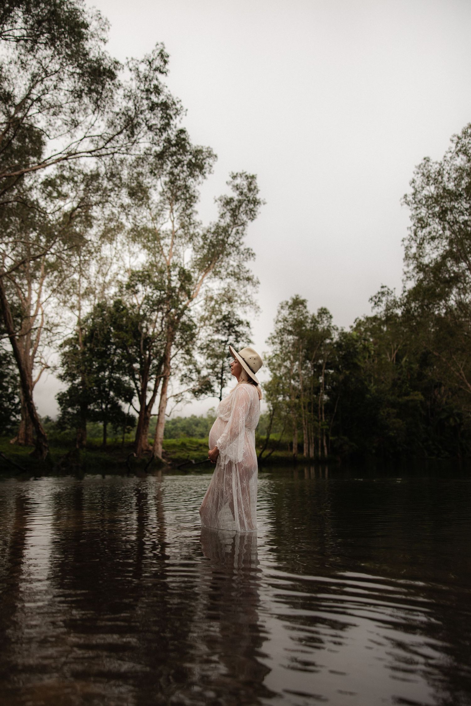 Ethereal maternity portrait of a woman in a flowing white dress standing in shallow water surrounded by trees.