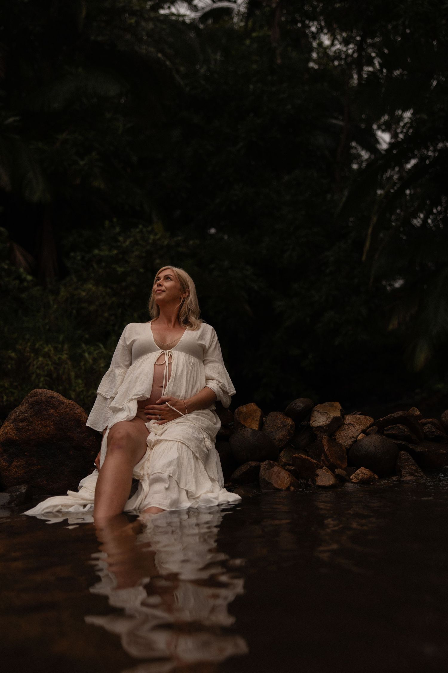 A person in a flowing white dress sits on rocks by a dark stream in a moody forest setting.