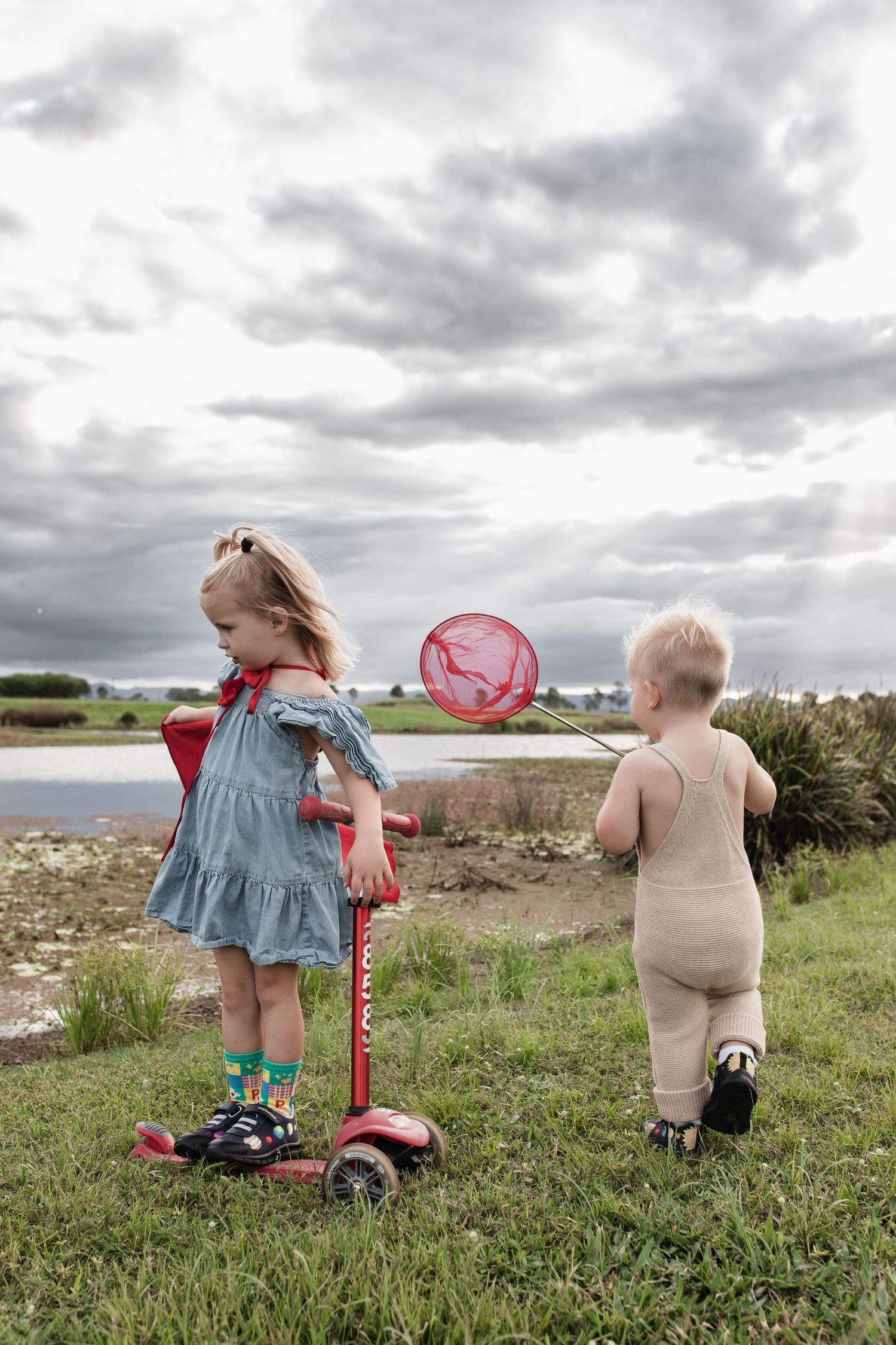 Two children exploring nature with a red scooter and butterfly net by the water.