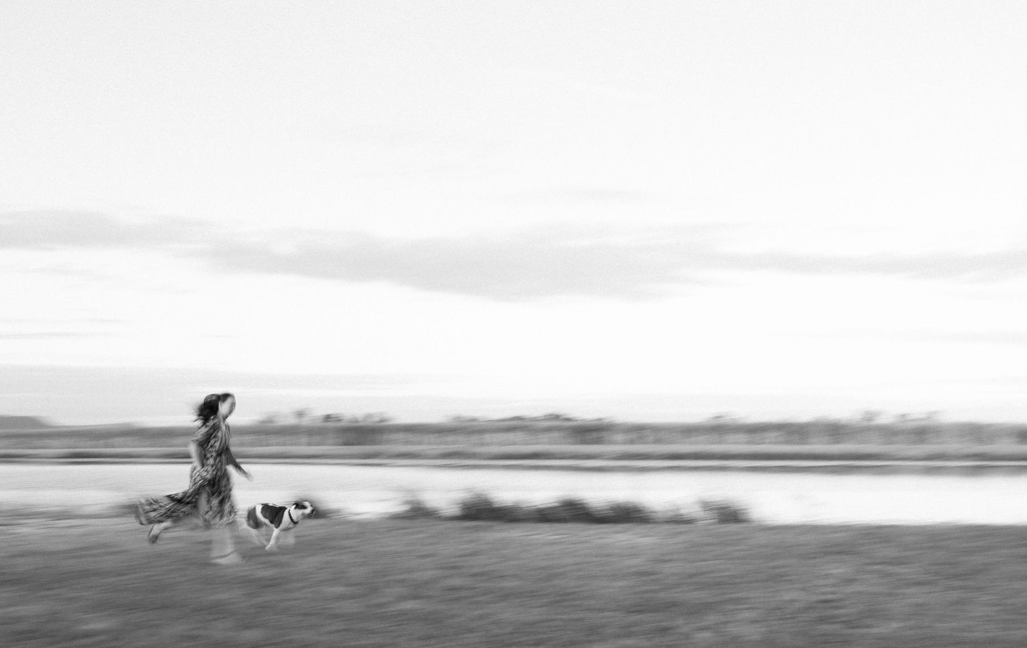 A silhouetted figure walks their dog along a misty shoreline at dawn in this serene black and white photograph.