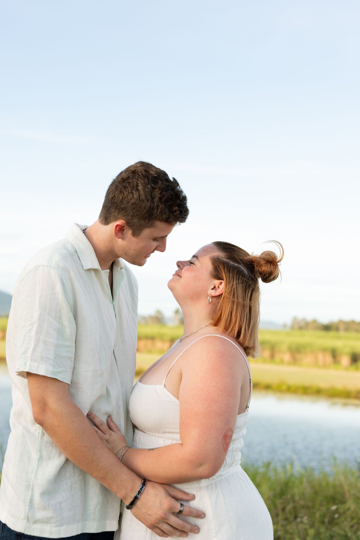 An affectionate couple embraces while standing near a lakeside on a sunny afternoon.