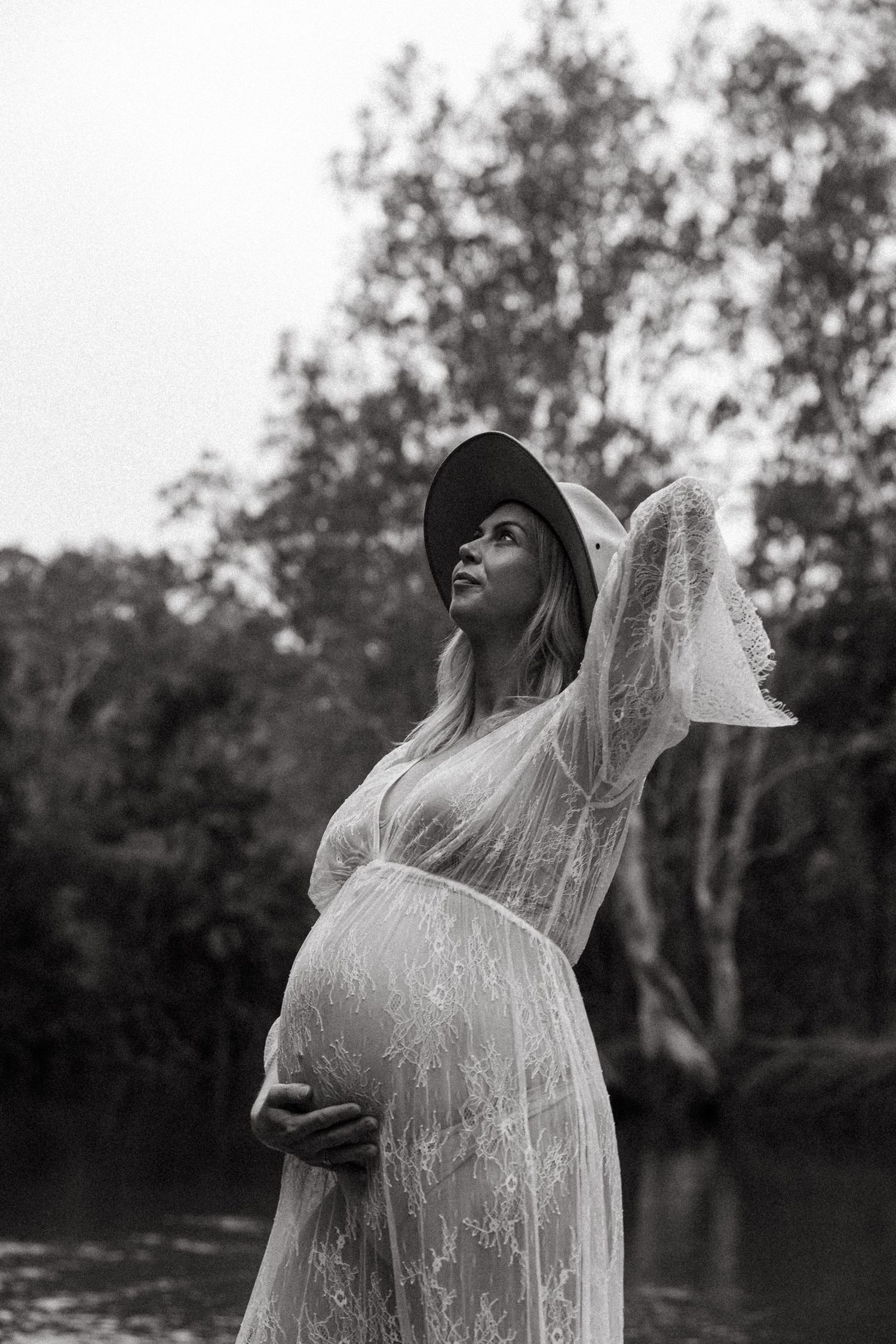 Black and white maternity photo of someone in a sheer lace dress and hat posing by a lake at dusk.