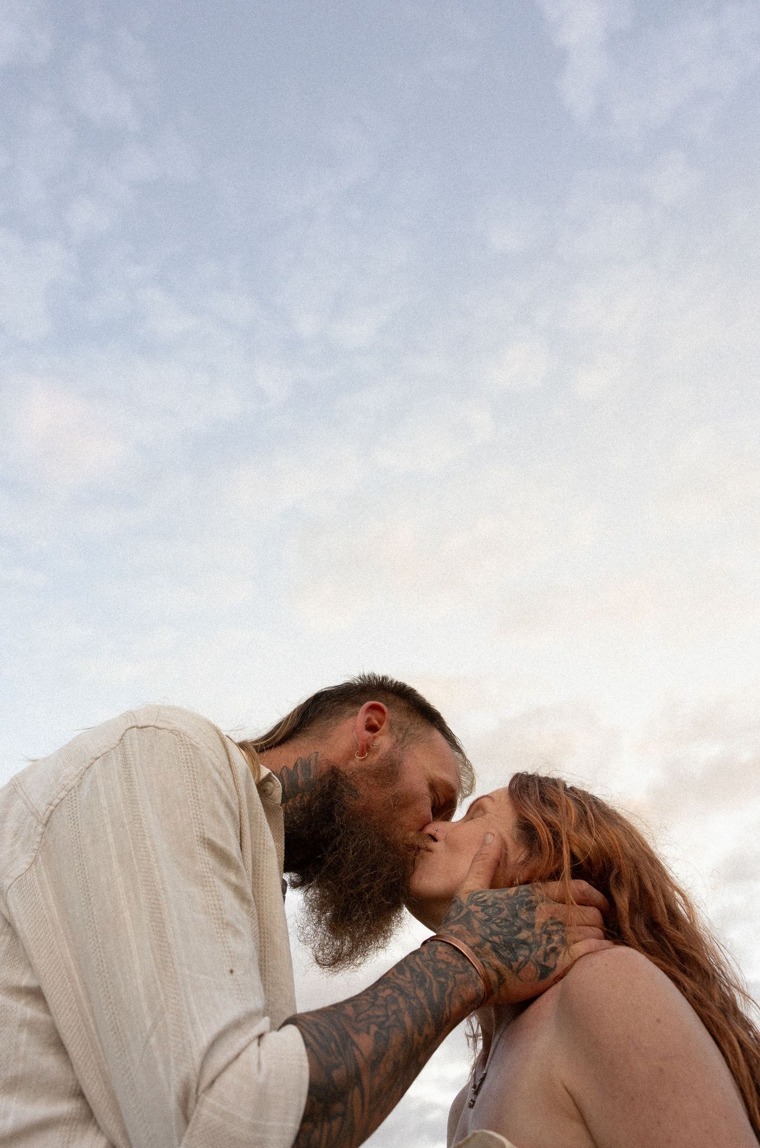 A couple sharing an intimate kiss against a cloudy sky background at sunset with visible tattoos and a full beard.