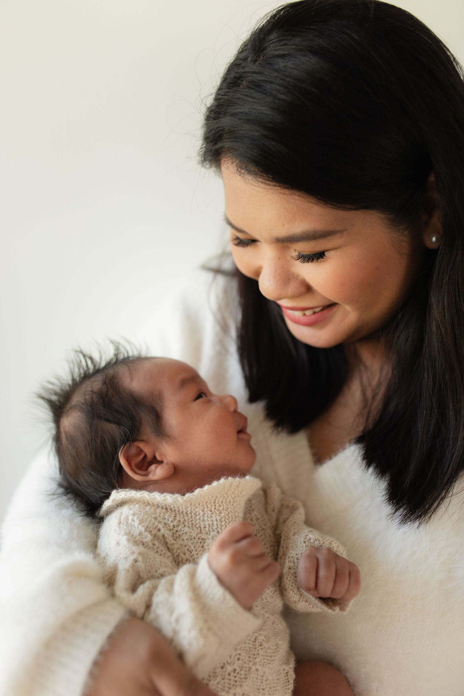 A tender moment between mother and newborn baby wearing a knit sweater against a bright white background.