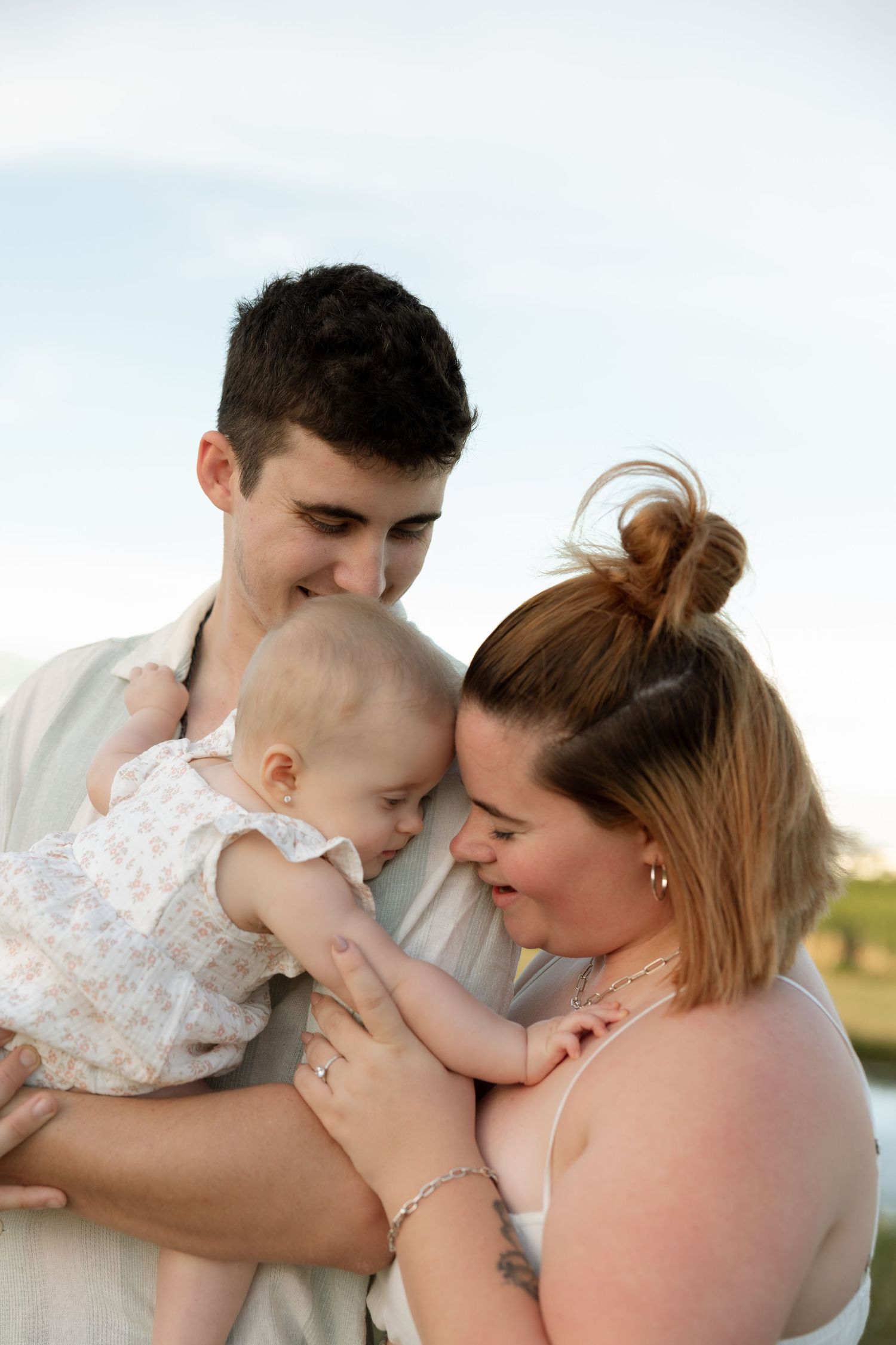 A tender family moment captured outdoors as parents share a loving interaction with their baby in white lace.