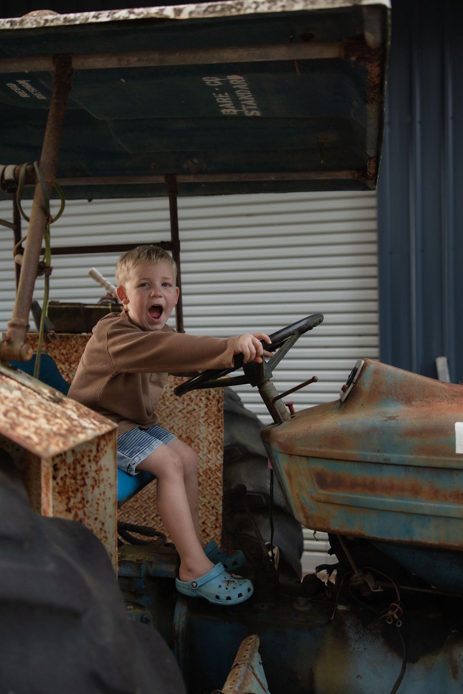 A young child sits excitedly at the steering wheel of an old rusty tractor, wearing a brown sweater and blue shoes.