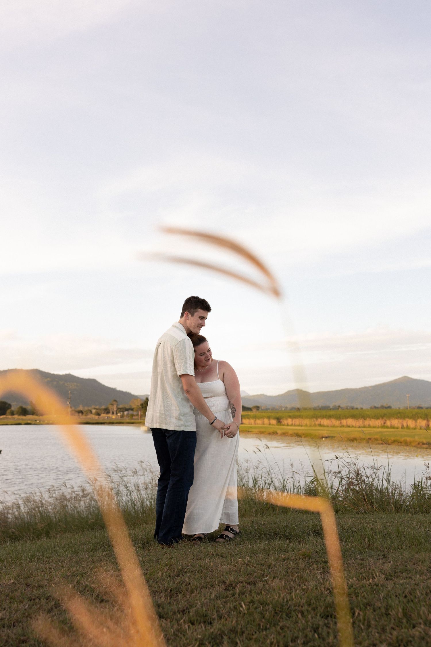 A couple shares an intimate moment at sunset by a tranquil lake with mountains in the background.