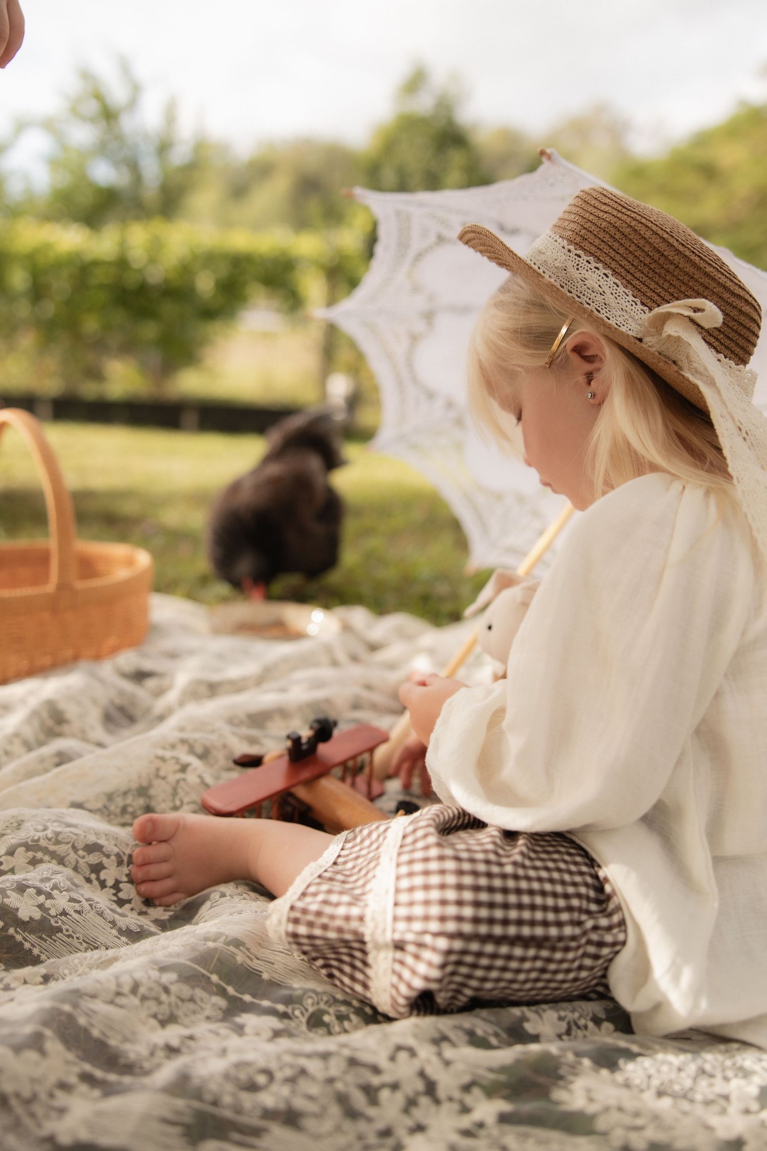 A straw hat adorned child sits on a picnic blanket with a parasol and black cat nearby on a sunny afternoon.