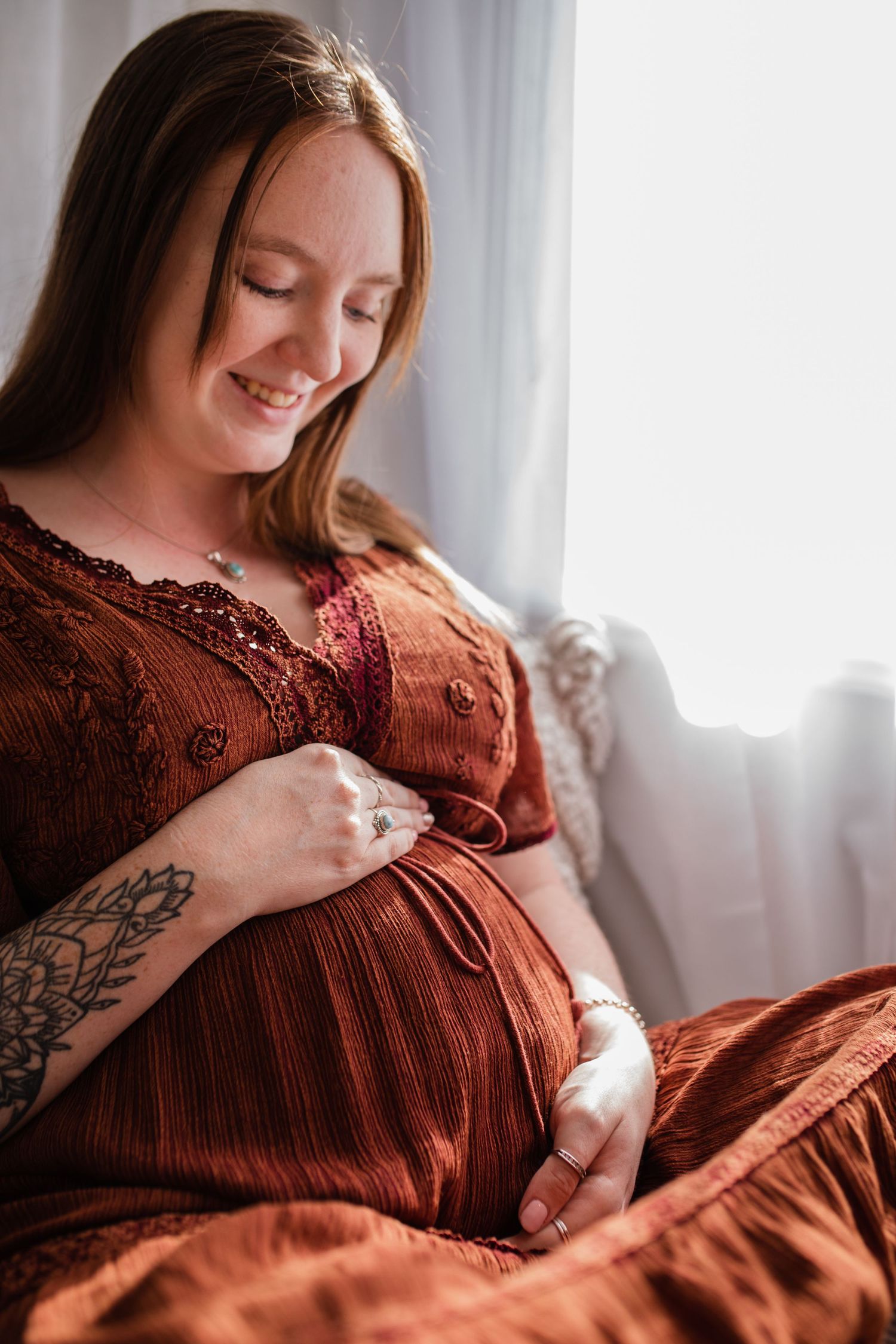Serene maternity portrait in rust dress by sunlit window.
