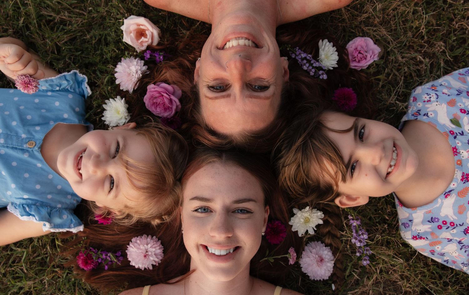 A group of people lie on grass in a circular formation with pink and white flowers placed around their heads.